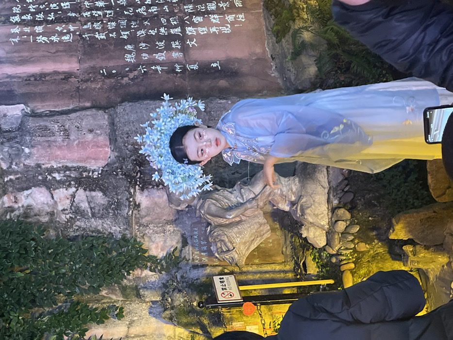 A young female, appearing to be of East Asian descent, is positioned centrally in a themed environment in Yuzhong District, China. She wears a light blue traditional-style Chinese dress with a translucent outer layer and a prominent, sky-blue floral headpiece. Her face features decorative glitter under her eyes and on her forehead. Her right arm is slightly extended, hand open, as she gazes towards her right.
Behind the subject is a rough, textured stone wall with Chinese characters carved into its surface. Green foliage cascades from the top left, partially obscuring the wall. A bronze-colored statue of a figure carrying a large basket is situated to the left and behind the female subject. To the far left, a black pole displays a "禁止喂食 NO FEEDING" sign, below which multiple orange heart-shaped ornaments hang from a chain. The ground is rocky, interspersed with small stones and green vegetation. The scene is illuminated by artificial lighting, casting warm yellow tones on the foliage and cooler light on the central figure. Portions of at least two other individuals are visible in the blurred foreground, with one holding a smartphone displaying a screen.