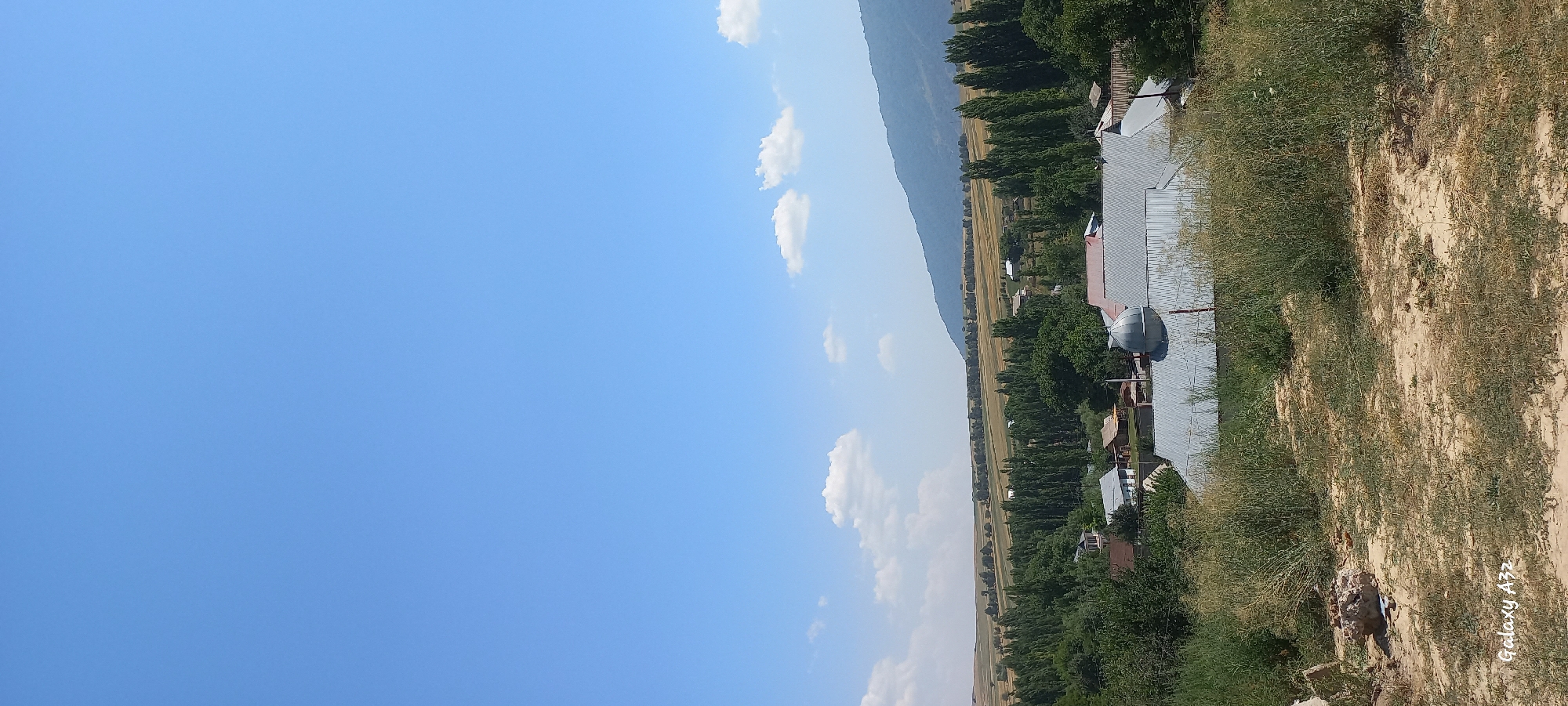 The image depicts a rural landscape near Joon‑Kunggöy, Kyrgyzstan, under a clear blue sky with scattered white clouds. In the foreground, a dry, sandy area is covered with sparse, dry grass and some larger stones. Beyond this, a cluster of buildings is visible, characterized by their grey, corrugated metal roofs. One building prominently features a domed roof structure. The village is nestled amongst numerous tall, slender trees, characteristic of poplar or cypress varieties, which form a dense line. Further in the background, a vast, open field, possibly agricultural land, stretches towards a gently sloping, tree-covered mountain range in the distance. The overall scene suggests a sunny, tranquil day in a mountainous region.