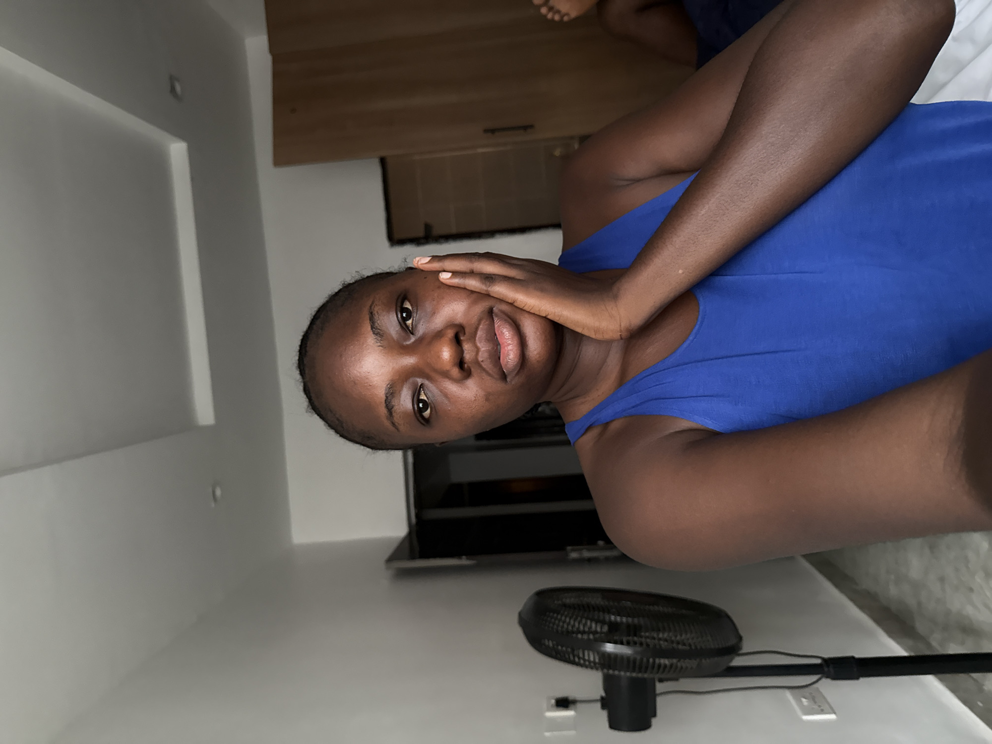 A woman with dark skin is indoors in a room in Iddo Tudunwada, Nigeria. She is wearing a blue sleeveless shirt and has her hand on her face. A black stand fan is visible in the foreground. A television and cabinet are in the background. The walls are white.