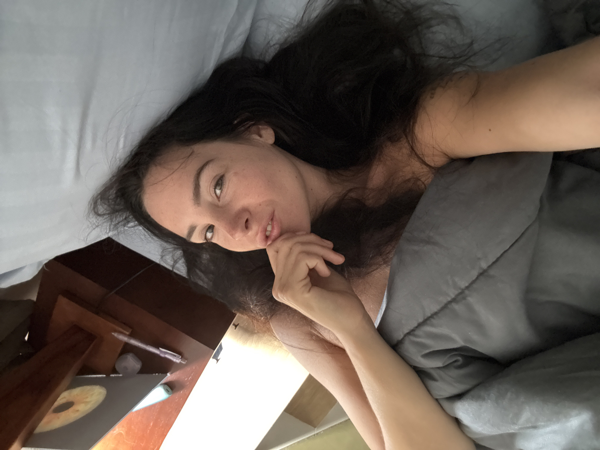 A woman with long, dark hair is lying in bed, looking at the camera. Her head is tilted to the side, and she is resting her chin on her hand. She appears to be awake and relaxed, with a slight smile on her face. The woman is wearing a light-colored top.
The scene is a bedroom, with a light blue sheet on the bed. A dark gray blanket is pulled up to her chest. To the left of the bed, a dark wooden desk is visible. On the desk, there is a laptop with a colorful design, a pen, and a small gray object that could be an eraser or a USB drive.
The lighting suggests it is daytime, likely morning or afternoon, as there is natural light coming from the side. The overall mood is intimate and casual, as if she is taking a selfie in bed. No specific time of day or weather is indicated beyond the daylight. The location context is North Kuta, Indonesia. There is no visible text in the image.