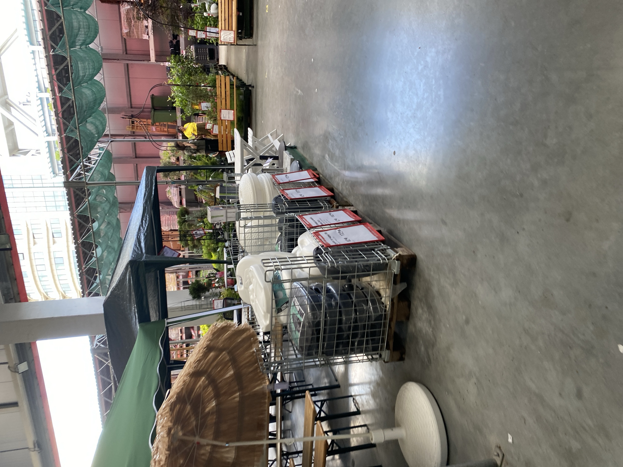 This is an interior shot of a large retail store, possibly a garden center or hardware store, in Varna, Bulgaria. The scene shows a variety of outdoor furniture and gardening supplies. In the foreground, a large, round, light-colored umbrella with a straw-like canopy is on a white base. To its right, a metal cage on a wooden pallet holds several large, white, plastic tubs or lids, and a stack of black cylindrical items, possibly trash cans or compost bins. Red and white price tags are attached to the front of the cage. In the mid-ground, there are wooden picnic tables and benches, along with a white foldable table and chairs. Further in the background, more shelving units with plants and gardening supplies are visible. Two women are present, one wearing a yellow shirt and the other a dark jacket, standing near some greenery. The overall environment is brightly lit, suggesting artificial lighting and possibly natural light coming from large openings. The floor is a smooth, gray concrete surface.