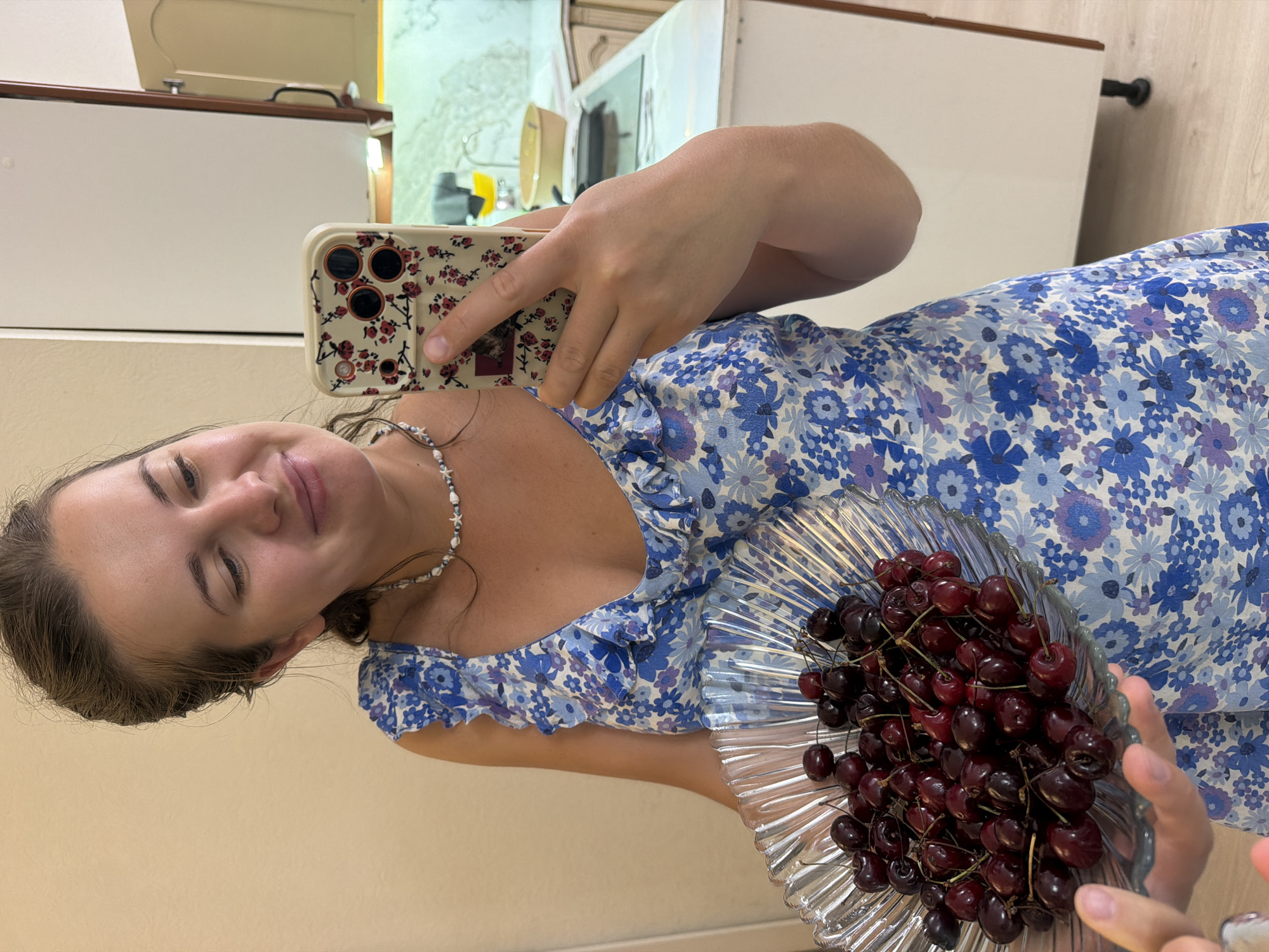 A young woman with dark hair is taking a selfie in what appears to be a kitchen. She is wearing a blue floral dress and a beaded necklace. In her right hand, she holds a clear glass bowl filled with dark red cherries. Her left hand is holding up a smartphone with a floral case, capturing the reflection of herself and the bowl of cherries. The background shows kitchen cabinets and countertops, with a hint of a tiled wall. The lighting suggests it could be daytime, and the overall mood seems cheerful and relaxed. The specific location is identified as Sochi, Russia.