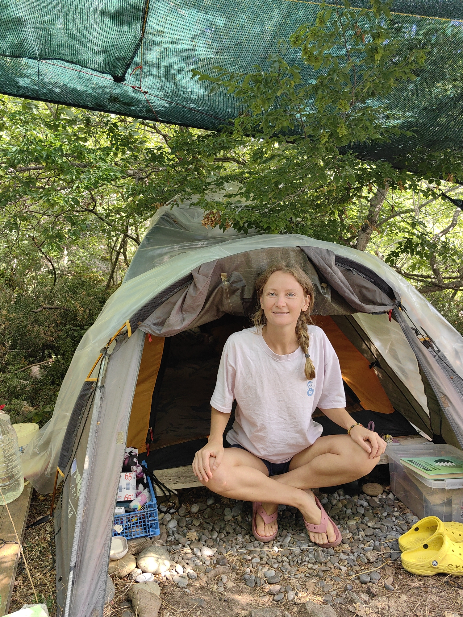 A blonde woman with a braided ponytail, wearing a pink t-shirt and dark shorts, sits cross-legged in front of a tent. The tent is made of grey and yellow fabric, with a clear plastic tarp draped over it. The ground around the tent is covered in gravel and pebbles. In the background, trees and foliage are visible, with a green mesh shade cloth stretched overhead. To the woman's right, a pair of yellow Crocs are placed on the ground. To her left, a blue plastic crate holds various items, including a bottle of water. The scene appears to be a campsite in Utios, Ukraine.