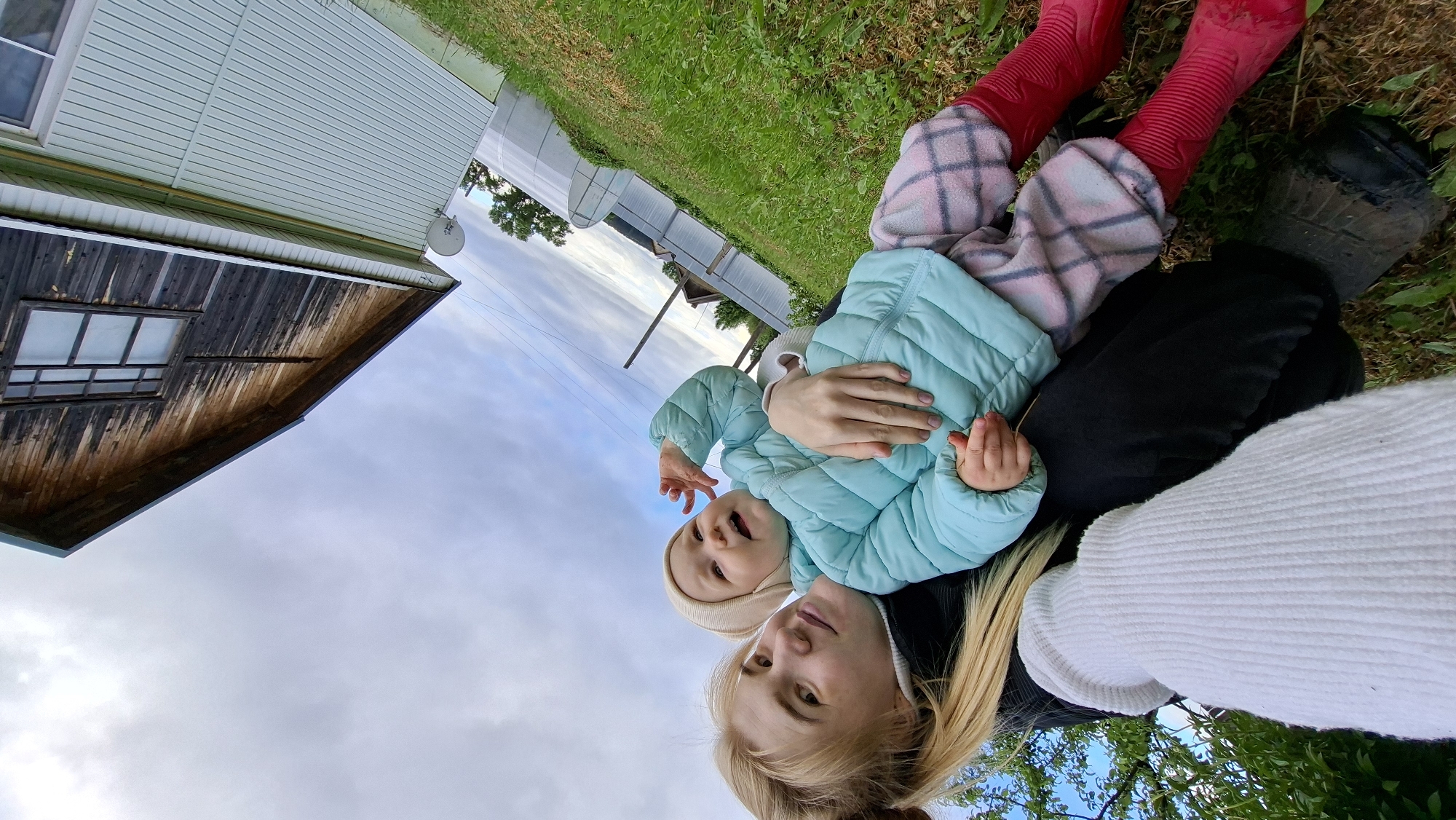 A mother is sitting in the grass in Большой Букор, Russia, cradling her baby. The baby is wearing a light blue jacket, red rubber boots, and plaid pants. The mother is wearing a white sweater. Behind them is a white building, a greenhouse, and a cloudy sky. A wooden building is in the foreground.