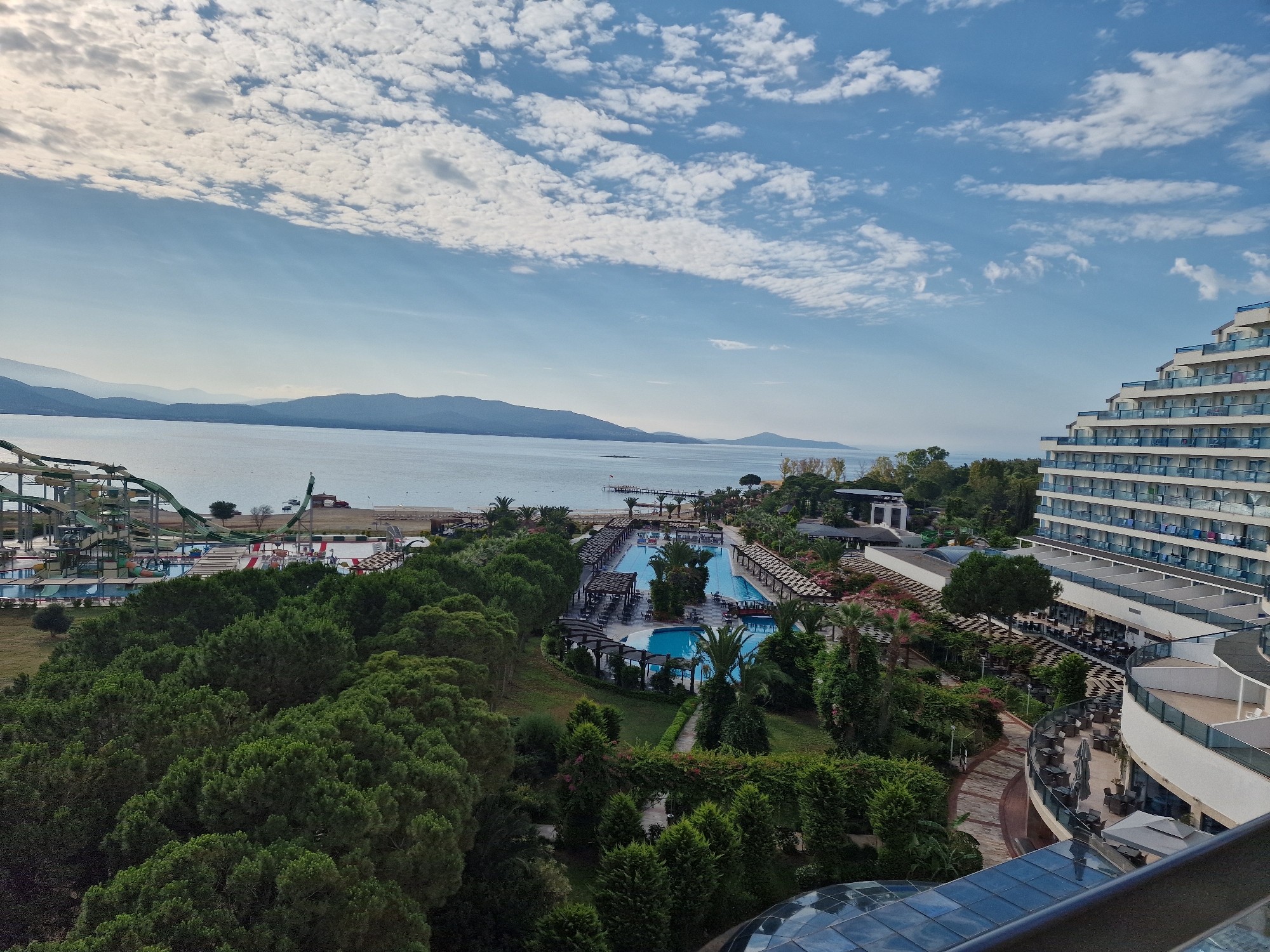 This is a wide shot of a resort area in Didim, Turkey, taken from a high vantage point, likely a hotel room. In the foreground, on the right, is a modern, multi-story hotel with a white facade and balconies with blue glass railings. The hotel's architecture is tiered, creating a stepped appearance. Below the hotel and to the left, a pathway with patterned brickwork winds through lush green trees and manicured lawns. Further into the scene, a large, rectangular swimming pool complex is visible, with lounge chairs and umbrellas arranged around it. Beyond the pool, a sandy beach meets a calm, blue body of water, likely the Aegean Sea. In the distance, hazy mountains form a picturesque backdrop. On the far left of the image, a section of a water park with green slides can be seen. The sky is a bright blue, dotted with fluffy white clouds. The overall impression is one of a vibrant, well-maintained resort on a sunny day.
