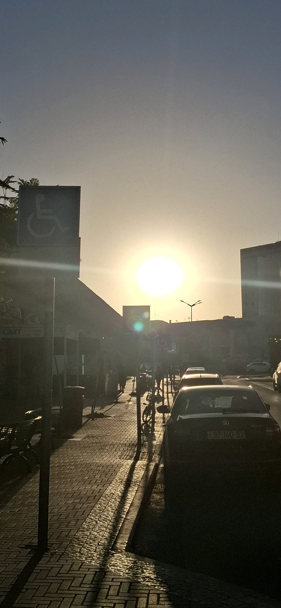 The image depicts a street scene in Be'er Sheva, Israel, during what appears to be sunset. The sun is low on the horizon, casting a bright, warm light and long shadows across the paved sidewalk and street. A prominent sign with the international symbol of accessibility for disabled individuals is visible on the left side of the frame, partially silhouetted against the bright sky.
Several cars are parked or driving along the street. One car in the foreground, a dark sedan with license plate "97 140 52", is clearly visible. In the background, buildings are present, one of which appears to be a modern structure. The sidewalk is constructed of interlocking bricks. A lamppost is visible in the middle ground, and a bicycle is propped up near the sidewalk. A few individuals are present, walking along the sidewalk, appearing as indistinct figures due to the backlighting and distance. The overall atmosphere suggests an urban environment at the end of the day, with the warm light creating a peaceful, yet active, mood. The sky transitions from a light blue near the top to a pale yellow and white near the horizon, characteristic of sunset or sunrise.