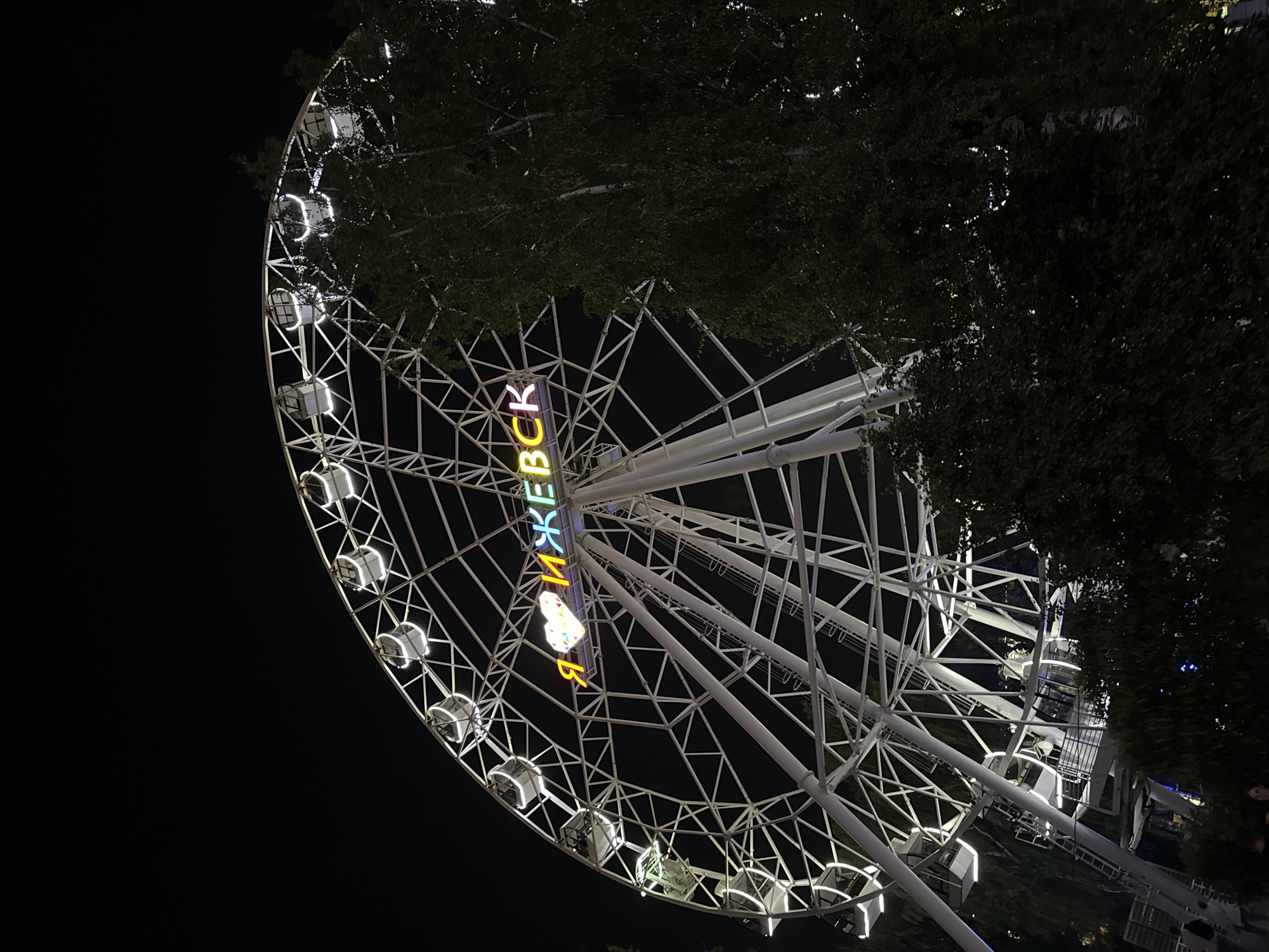 An illuminated Ferris wheel stands against a dark night sky in Izhevsk, Russia. The wheel is decorated with neon lights that spell out "Я ЛЮБЛЮ ИЖЕВСК" which translates to "I LOVE IZHEVSK". The letters are a mix of yellow, pink, and blue. The Ferris wheel has white gondolas, some of which are illuminated from within. Trees with dark green foliage are visible in the foreground and partially obscuring the right side of the Ferris wheel. The lower part of the image shows a few people standing near the base of the Ferris wheel, their features not clearly discernible due to the low light conditions. The scene appears to be an amusement park or public space at night.