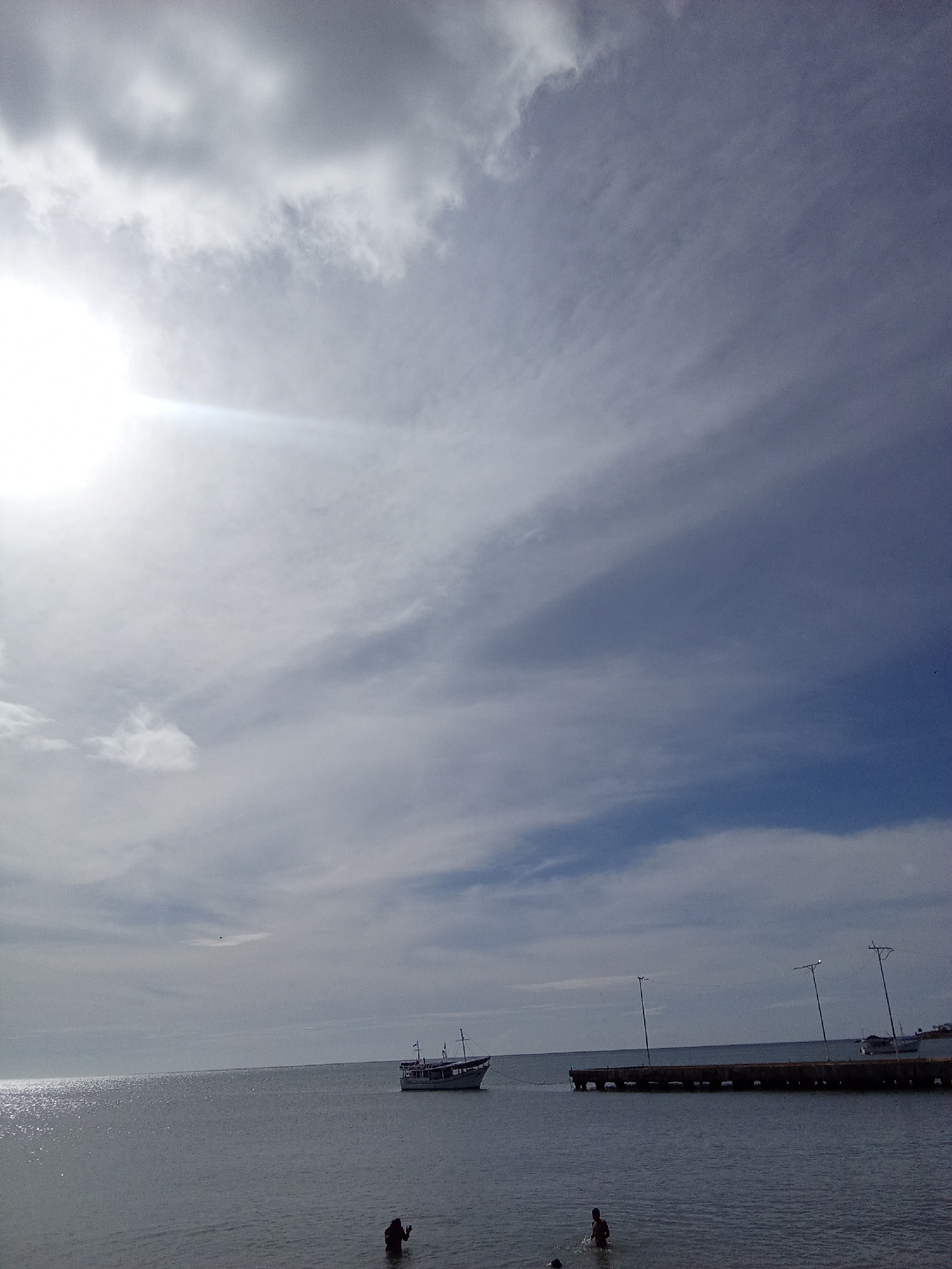 The image shows a waterfront scene in Juan Griego, Venezuela. A body of water, likely a bay or ocean, stretches from the foreground to the horizon. Above, the sky is filled with clouds. The sun shines brightly in the upper left, creating a glare. In the water near the lower center, two people are partially submerged. A boat is anchored in the distance, slightly to the left of center. A pier extends into the water on the right side of the frame, featuring several light posts.