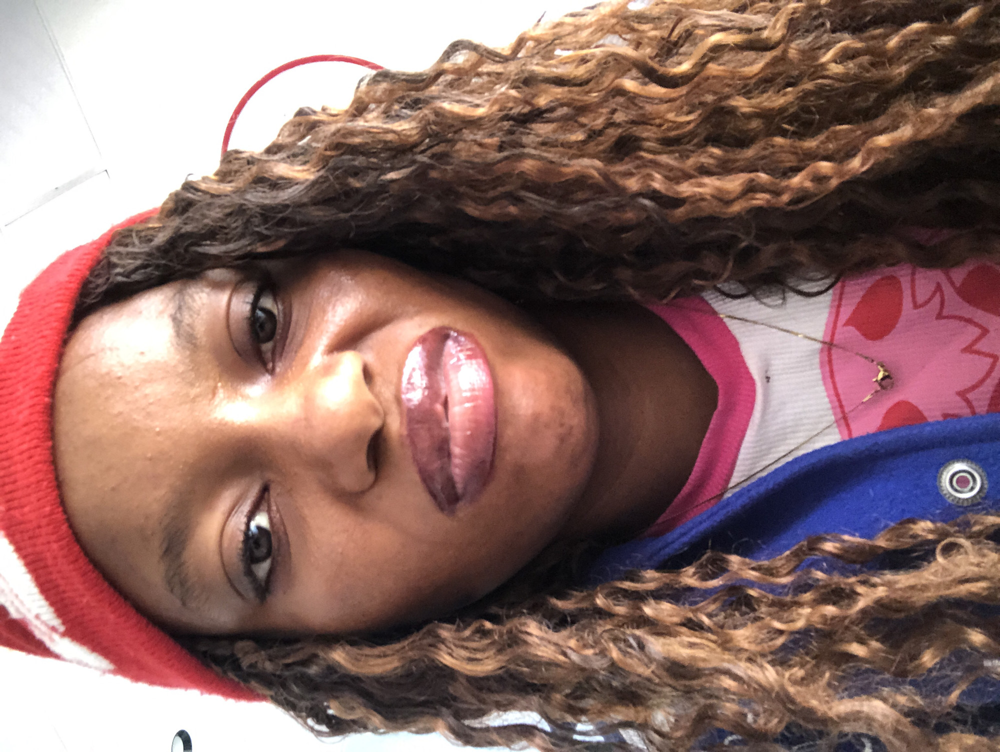 A medium close-up shot from below depicts a Black female wearing a red and white striped beanie. The woman's curly brown hair frames her face. Her lips are covered in glossy makeup. She is wearing a shirt with a pink collar and a pink print, partially covered by a blue garment. The background appears to be a neutral indoor setting. There are no indications the image was taken in Pindiga, Nigeria.
