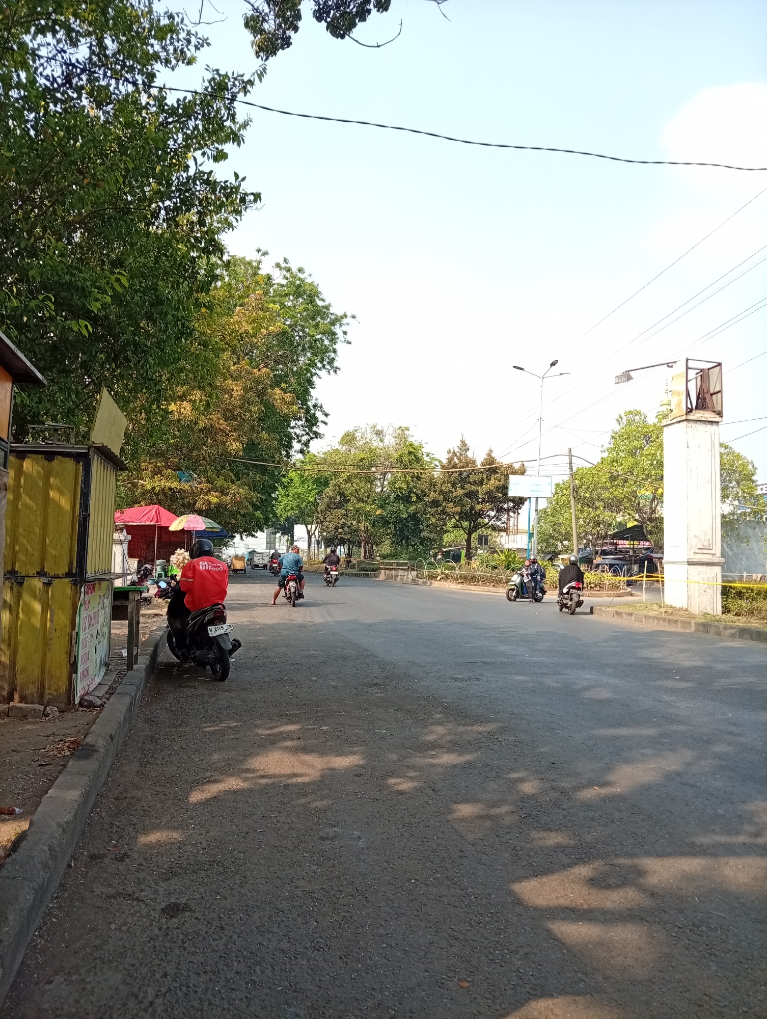 The image depicts a street scene in Sumokali, Indonesia, bathed in daylight. A paved road occupies the foreground, with a concrete curb and sidewalk on the left, lined with a yellow and white structure resembling a food stall or kiosk. Several individuals are present, all on motorbikes. In the immediate foreground, a person wearing a red jacket with a "Shopee Food" logo is seated on a black motorbike. Further down the road, two individuals are seen riding motorbikes. The right side of the image features a large, white, pillar-like structure and a metal pole with a street lamp. Trees with green foliage line the sides of the road, and power lines are visible against a clear, light blue sky. The overall environment suggests an urban or suburban setting with moderate activity.