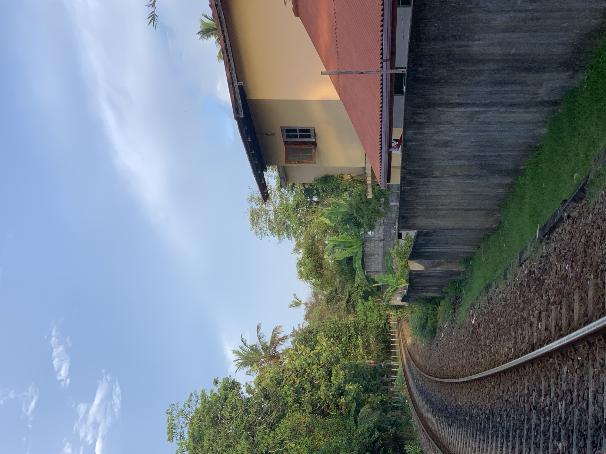 A low-angle, outdoor, vertical shot captures a railway track curving to the left, bordered by lush green trees and foliage on the left side. A grey concrete wall runs along the right side of the track, separating it from a property. A yellow house with a red-tiled roof is visible behind the wall, with an open wooden window. The sky above is a clear blue with scattered white clouds. The scene appears to be located in Kabalana, Sri Lanka, given the tropical vegetation and architecture. There are no people or animals visible in the image. The overall atmosphere is serene and suggests a quiet afternoon in a rural or suburban setting.