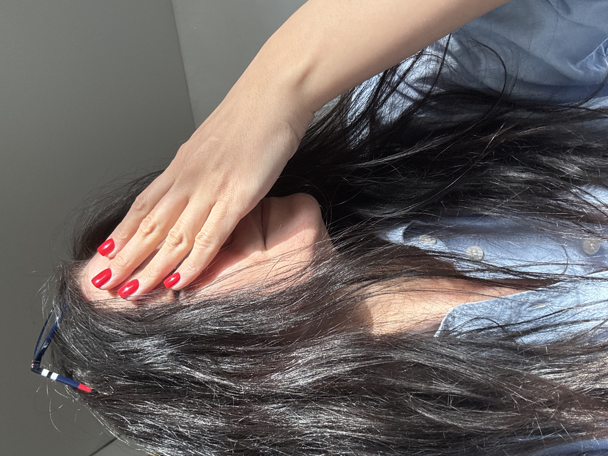 A person with long, dark hair is covering their face with their right hand, obscuring their eyes. The person is wearing a light blue collared shirt. The fingernails on their right hand are painted bright red. A pair of glasses with a dark blue and white frame is visible in the lower-left corner. The background appears to be a light grey or white wall. There is no indication this image was taken in the city of Chaikovsky, Russia.