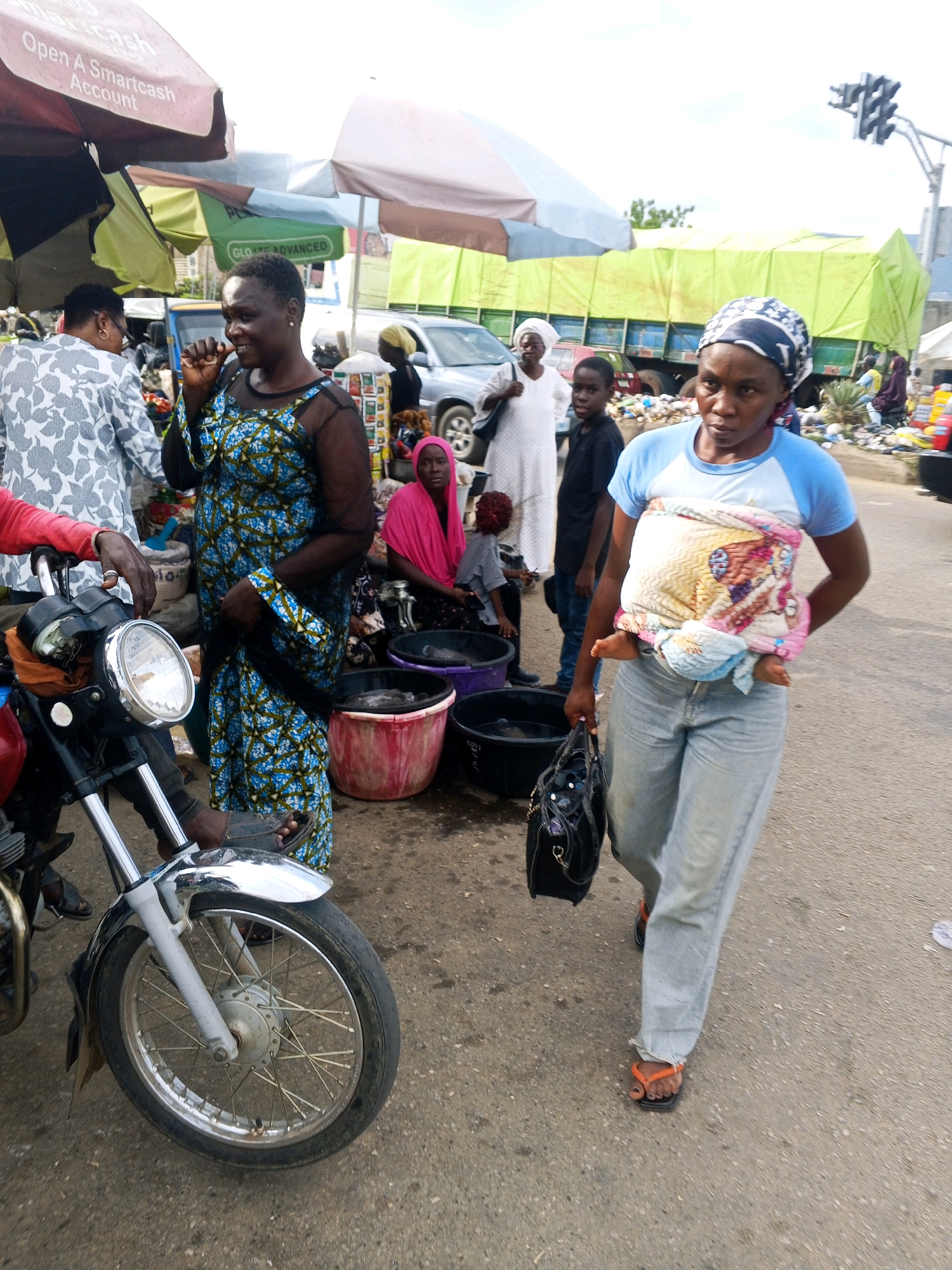 The image depicts a bustling street scene in Lokoja, Nigeria. A woman with a headscarf wrapped in a colorful fabric is carrying a baby strapped to her front. She wears a light blue t-shirt and light-wash jeans. Beside her, a woman in a blue and green patterned dress is looking towards the left of the frame, her hand near her mouth as if in thought. To the far left, a person in a white patterned shirt stands by a motorcycle. In the background, a large green tarp covers a truck, and a traffic light is visible against a bright sky, suggesting daytime.
The scene appears to be a market or roadside stall area, with various buckets and containers on the ground. One pink bucket and two black buckets are prominent in the foreground, possibly containing goods or water. The ground is paved with asphalt and shows signs of wear. The overall impression is one of everyday life and commerce. The presence of the motorcycle, the various people in different attire, and the market-like setup indicate a public space where people are going about their daily activities. The weather appears to be clear and bright. There is no visible text in the image.
