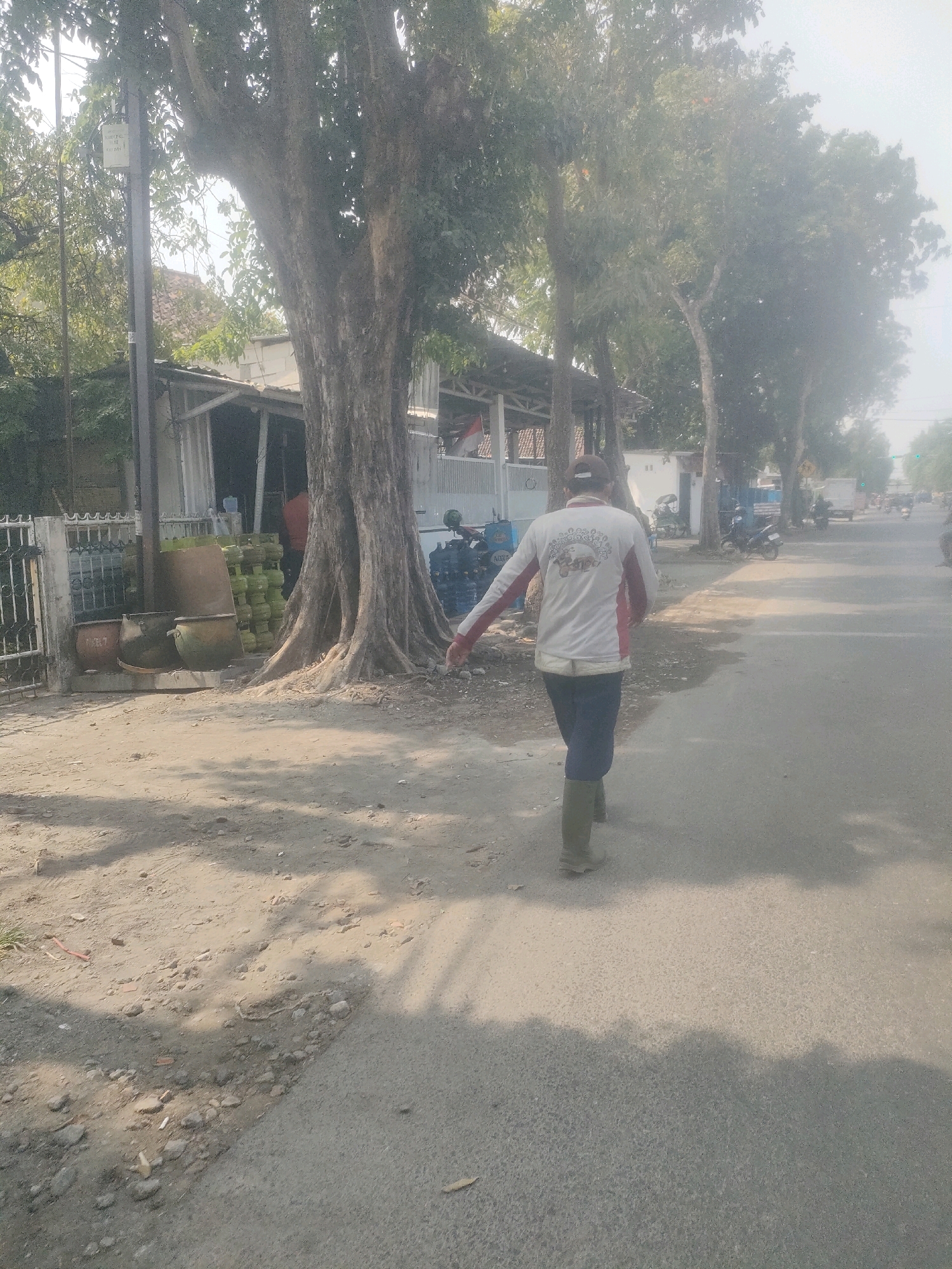 A man wearing a white shirt with red accents, dark pants, and green boots walks on the right side of a paved road in an outdoor urban environment. He is walking away from the camera and towards the left of the frame. His right arm is extended outwards, with his hand open. The road is asphalt and appears to be a two-lane street. To the left of the road, there is a dirt path leading to a building with a covered porch. In front of the building, several large earthenware pots are visible, along with stacks of green gas cylinders. A large tree with a thick trunk is situated on the edge of the dirt path, casting shadows on the ground. Behind the building, more trees and structures can be seen. Further down the road, several motorcycles and a white truck are present, suggesting it is a moderately busy street. The sky is bright and hazy, indicating a sunny day. The scene appears to be in Surabaya, Indonesia, given the presence of gas cylinders commonly used for household fuel, and the architectural style.