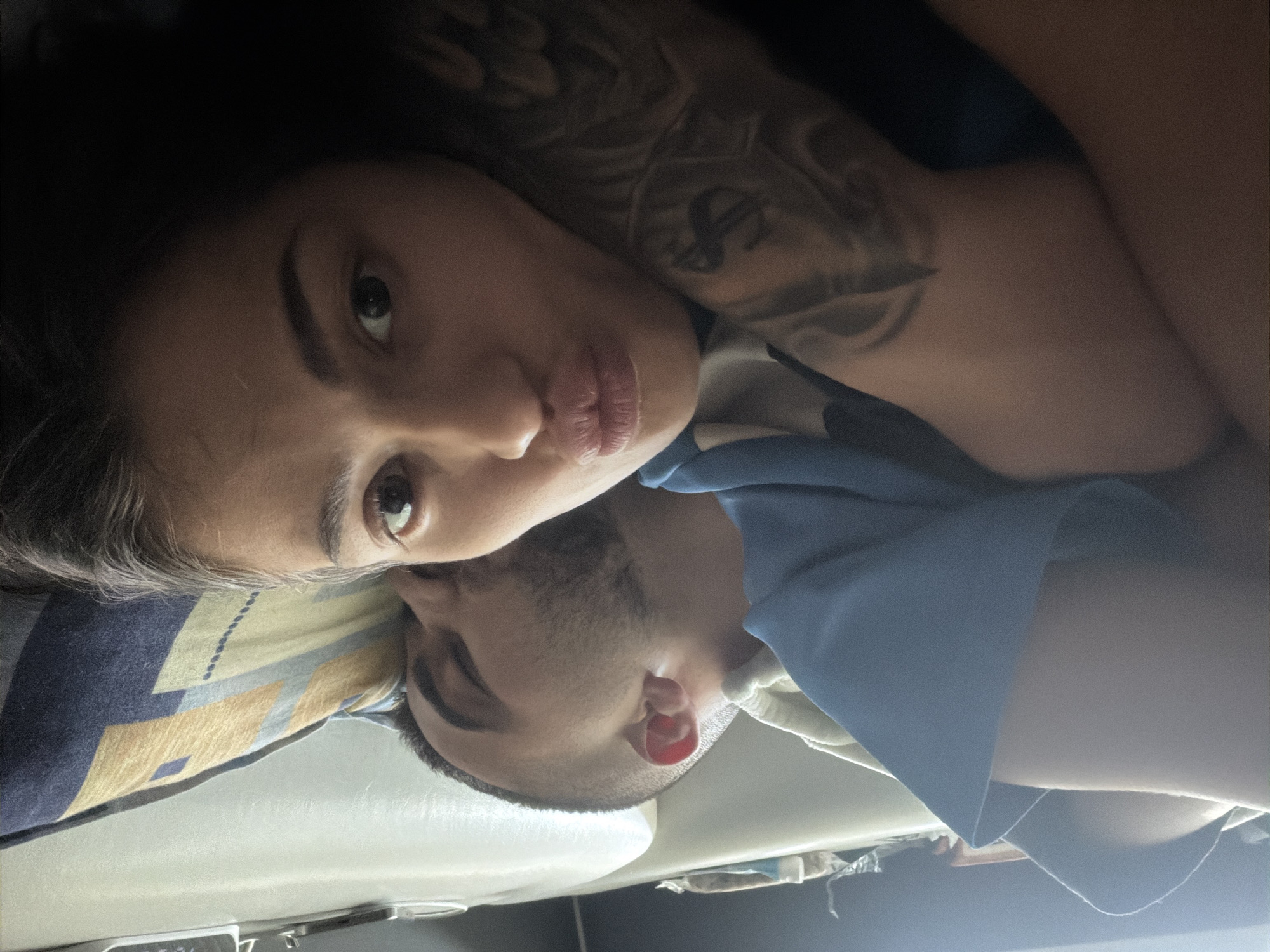 A selfie-style photograph shows a woman and a man lying down together. The woman is positioned closer to the camera, with her face angled towards the lens. She has dark hair, a light complexion, and prominent lips, and she appears to be making a kissing face. Visible on her upper chest and shoulder is a tattoo.
The man is lying behind her, with his face angled toward the camera but slightly obscured by her. He has dark hair that is cut short, and a trimmed beard.
They appear to be lying on a bed with a patterned pillow. There is a blue garment draped over them.
The location of the photo is Unknown, Unknown.