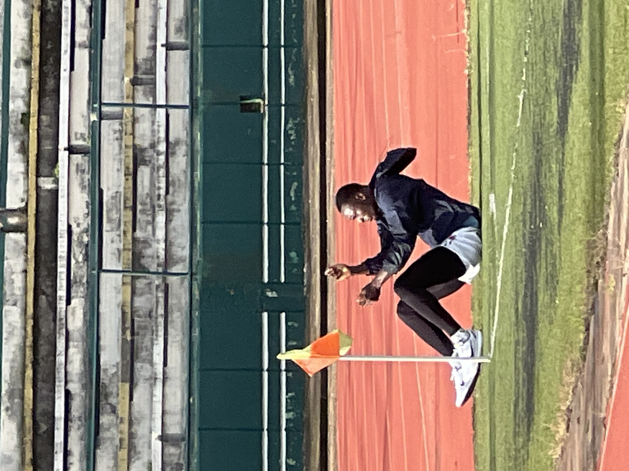A man is seated on the grass next to a running track, near a corner flag. The man is wearing a dark jacket, white shorts, black leggings, and white athletic shoes. He is looking to his left and has his hands raised. The running track is orange and there are stadium seats in the background. The image appears to have been taken in New Stadium, Nigeria.