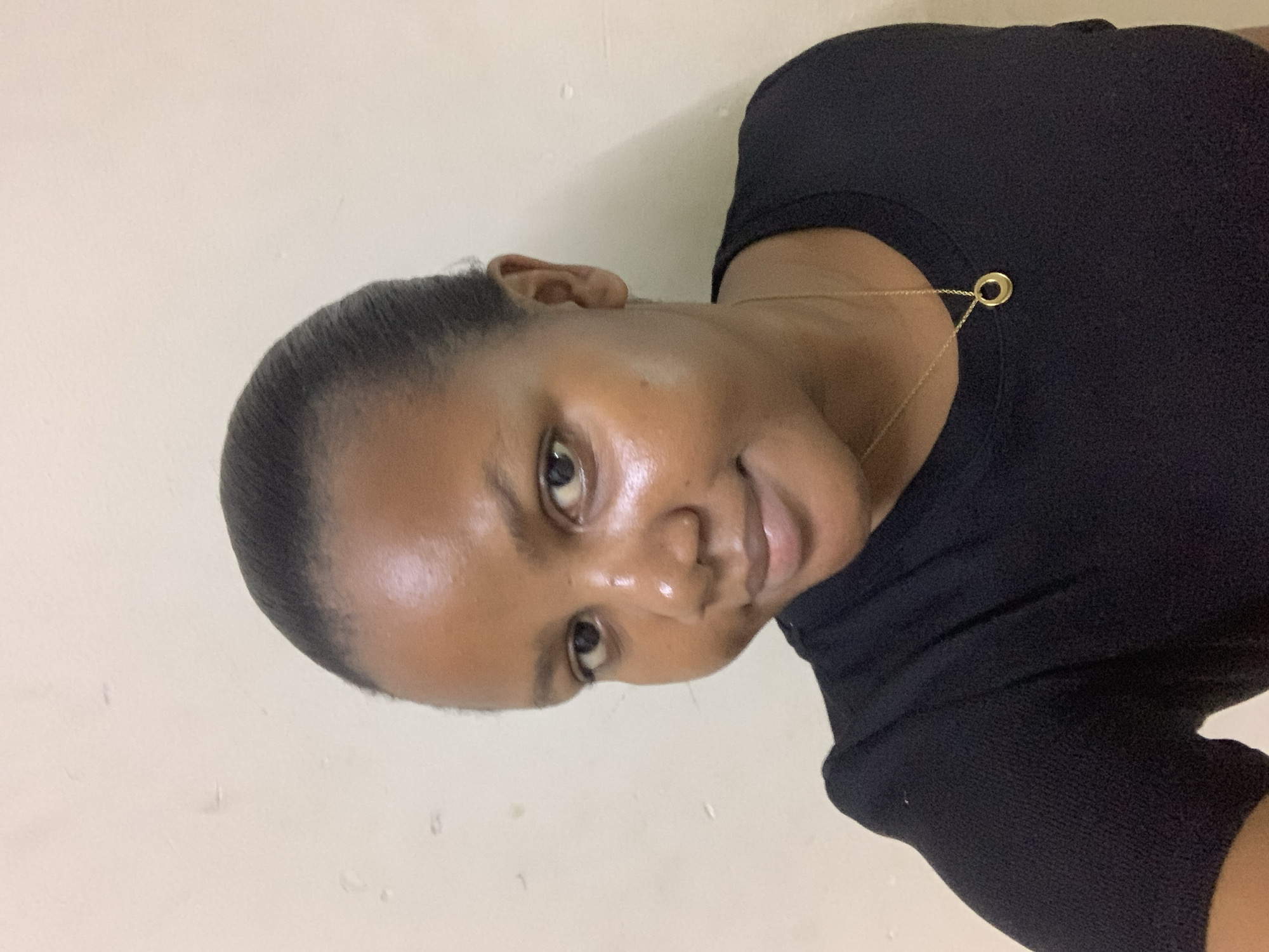A medium close-up shot depicts a woman with dark skin, short, dark hair, wearing a black top and a delicate gold necklace with a circular pendant. She is facing the camera with a slight smile. The background is a plain, off-white wall. The photo is lit softly, emphasizing the smoothness of her skin. There is no information available to confirm the location is in the city of Dar es-Salaam, Tanzania.