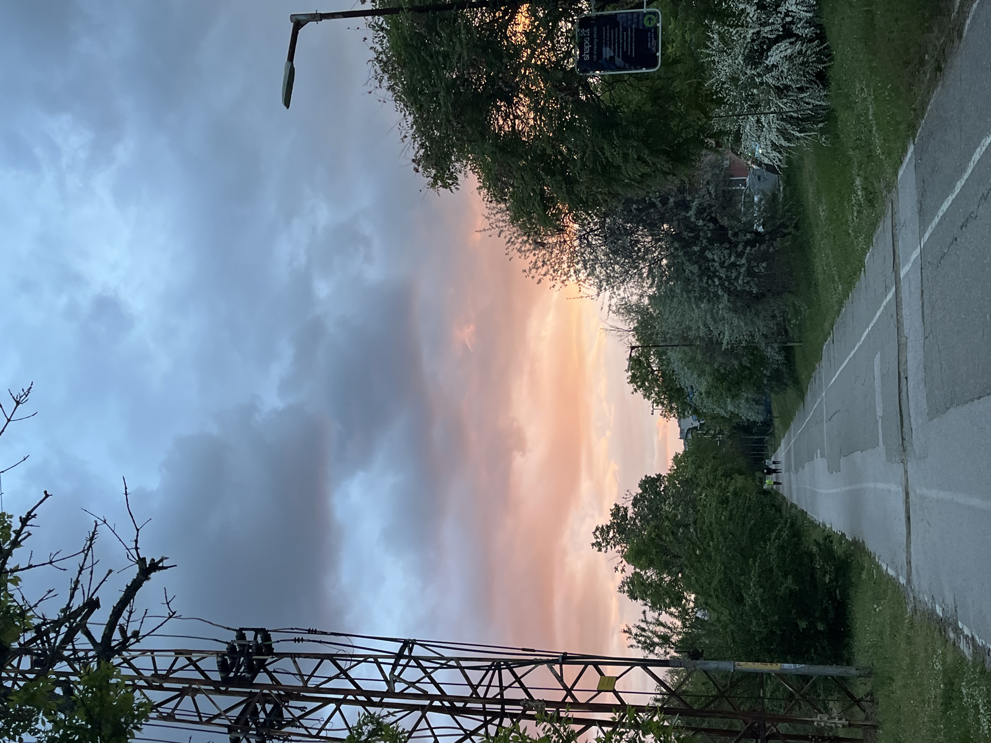 The image depicts an outdoor scene at either sunrise or sunset, characterized by a striking sky and a path lined with vegetation. The sky transitions from dark blue-grey clouds on the left to warm orange and pink hues on the right, illuminated by the low sun.
A paved road or path, marked with white lines, extends from the foreground into the distance on the right side of the frame. Lush green trees and bushes, some displaying white blossoms, line both sides of the path. A modern streetlight is visible in the upper left corner, extending over the path.
On the right side, within the trees, a black and white rectangular sign is visible. The text "shape.it" and "roll shaping studio" are clearly discernible, along with smaller print and a graphic element, possibly a QR code. In the mid-ground, three individuals are seen walking away from the viewer along the path. Two of them are wearing bright, possibly reflective, vests.
The lower left portion of the image features a prominent metallic lattice structure, likely associated with power lines or a railway, complete with visible wires and insulators. The scene combines natural elements with infrastructure, suggesting a park or a recreational path within an urban or suburban setting, under the dramatic light of dawn or dusk. The location context is Blagoevgrad, Bulgaria.