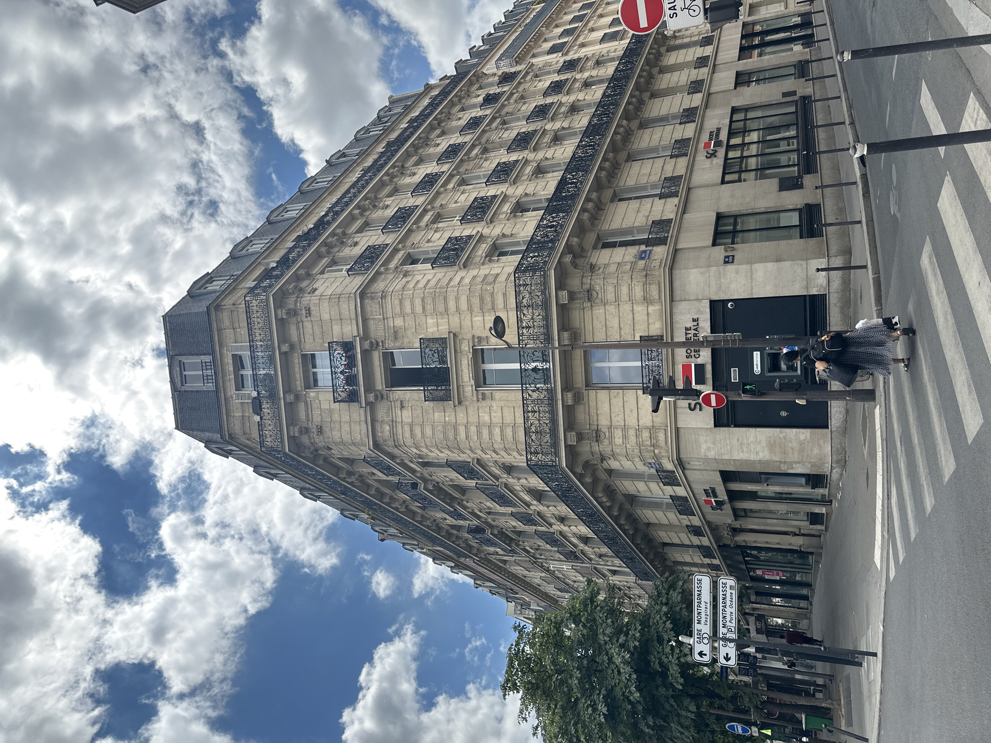The image shows a wide, upward-angle view of a light-colored stone building with a mansard roof on a street in Paris, France. The building has multiple floors with numerous windows and ornate black wrought-iron balconies. The street level features storefronts, including a Société Générale bank with its distinctive logo. Pedestrians are visible on the sidewalk and crossing a street at a crosswalk. Traffic signs are present, including one indicating "GARE MONTPARNASSE Vaugirard" and "GARE MONTPARNASSE Porte Océane," suggesting the building is located near Montparnasse station. The sky above is partly cloudy with blue sky peeking through. A green tree is seen on the left side of the frame. The overall scene depicts a typical Parisian street corner with a mix of residential and commercial architecture.