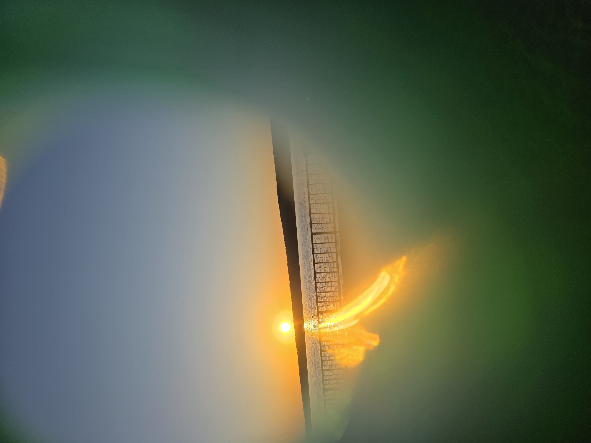 The image appears to capture a sunset over the Kama River in Chaikovsky, Russia. The sun is reflecting off of the water's surface, creating a bright glow. There is a vertical structure (possibly a bridge support or pier) intersecting the horizon, with a ladder attached to its side. The image has a distorted, almost lens-flare effect, creating a blurry green and blue frame around the central scene. The overall composition is abstract and emphasizes the light and color of the sunset.