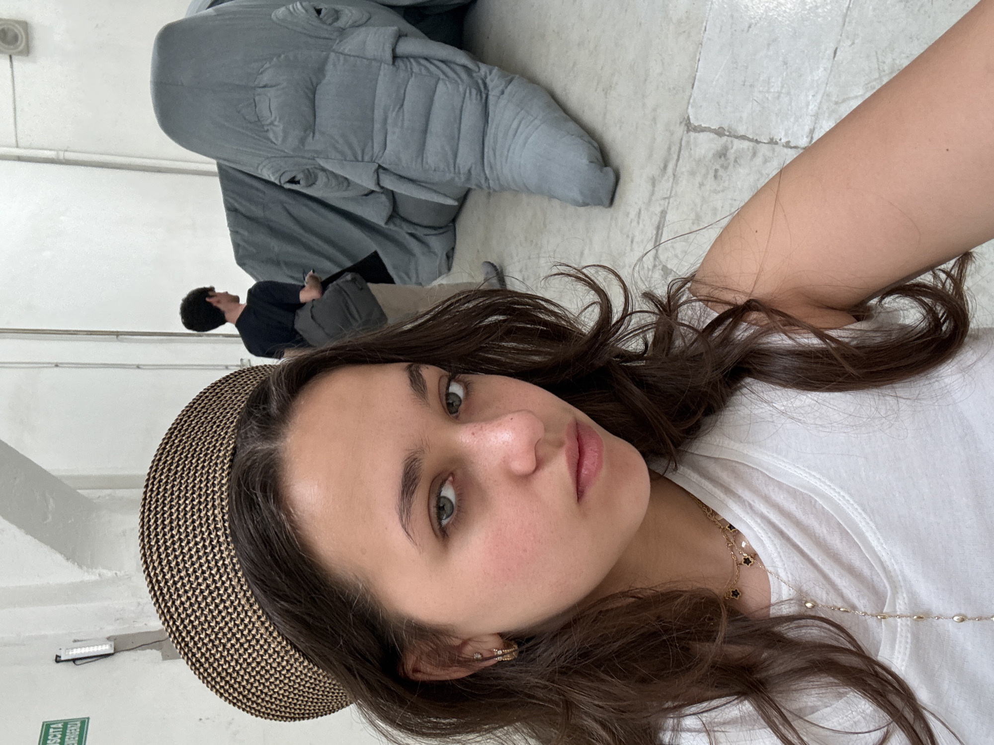 This rotated selfie captures a young woman in an indoor setting, likely an art gallery or museum in Milan, Italy. The main subject, a woman with medium-long dark wavy hair, fair skin, and light-colored eyes, looks directly at the camera with a neutral expression. She wears a white t-shirt, subtle gold necklaces (one with black clover-like charms), and small hoop earrings. A portion of a patterned straw or woven hat is visible on her head.
In the background, a man with dark hair, wearing a dark t-shirt and grey pants, is seen looking upwards or to the side, seemingly observing an exhibit. Dominating a significant part of the background is a large, grey, soft-looking, and amorphous object, which appears to be an artistic installation or sculpture.
The setting features light-colored, possibly concrete or tiled, flooring and plain white walls. Visible on the wall are a white pipe and a small light fixture. A green sign, partially visible in the bottom left corner (when viewed upright), clearly displays the Italian text "USCITA DI EMERGENZA," which translates to "EMERGENCY EXIT," corroborating the location context of Milan, Italy. The overall lighting suggests it is daytime indoors.