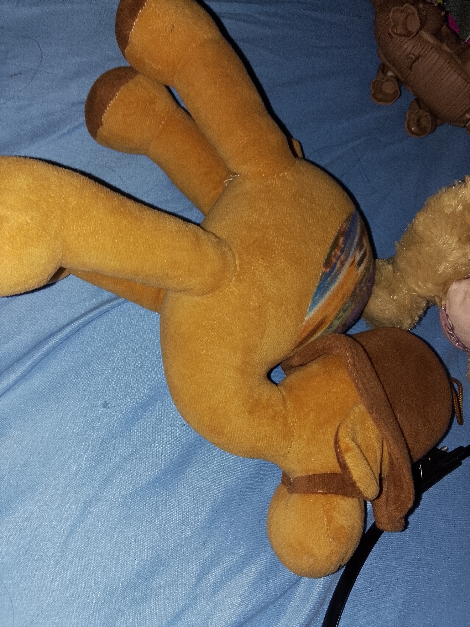 The image displays a collection of plush and plastic toys resting on a light blue fabric surface, likely a bedsheet or blanket, in an indoor setting within Davao City, Philippines.
The most prominent subject is a medium-sized plush horse toy, predominantly tan or light brown, lying on its left side. Its four legs are extended, and it has darker brown hooves. A colorful, abstract pattern resembling a landscape or tribal design is visible on its right flank. The horse also features a darker brown mane or harness around its head and neck area, along with a strap around its muzzle.
To the right of the plush horse, a smaller, furry plush toy, light brown with hints of pink, is partially visible. Further to the upper right, a textured, brown plastic toy, possibly a dinosaur or another animal with ridged skin and distinct legs, is also seen. A black cord extends from the bottom right corner of the frame, passing under the head of the plush horse.
There is no specific activity or event taking place; the toys appear to be casually arranged. The lighting suggests an indoor environment, but the time of day and specific weather conditions are not discernible. No visible text is present on any of the objects or in the background.