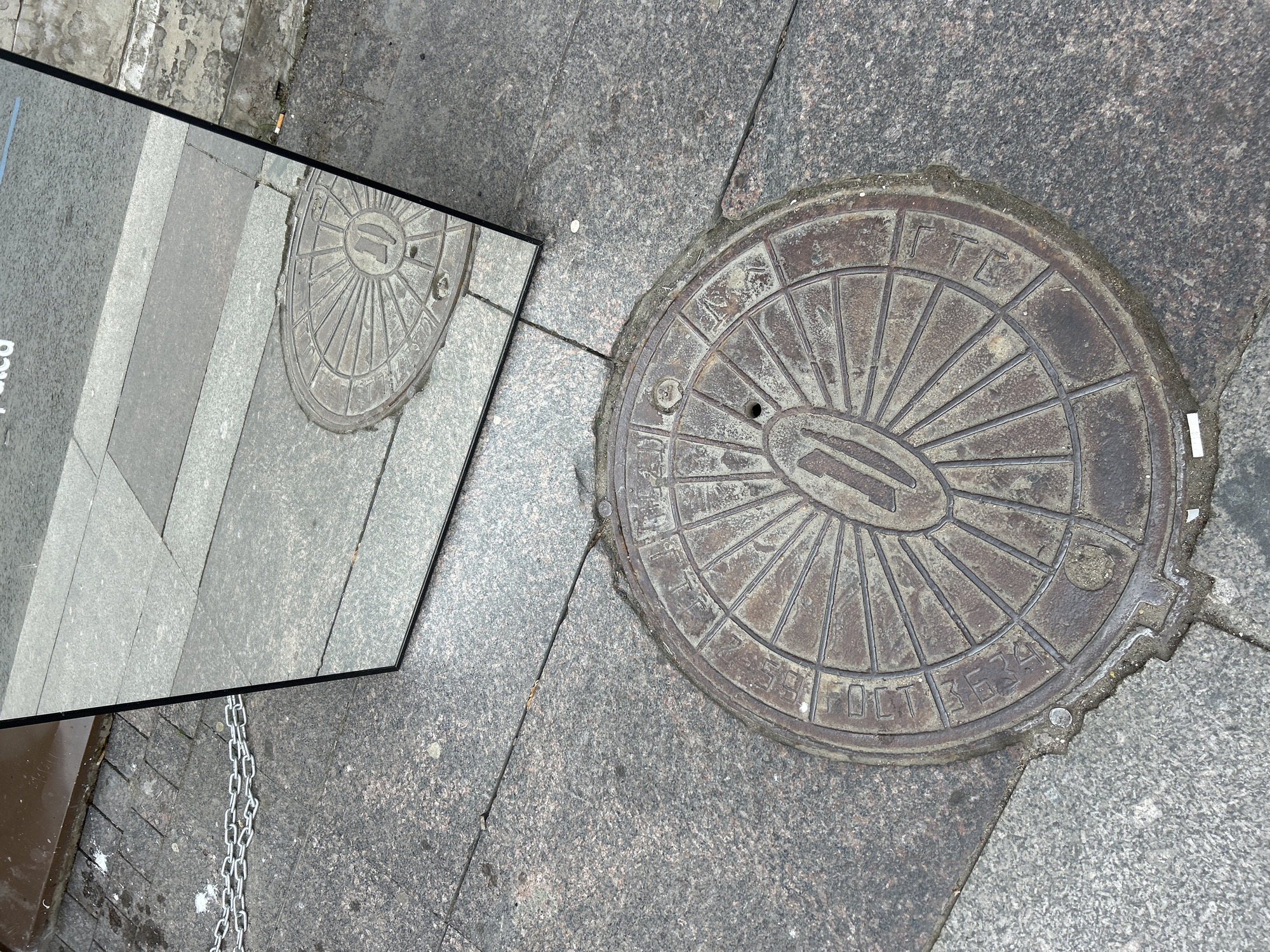 An outdoor, eye-level shot captures a ground-level urban scene featuring a circular, rusted metal manhole cover and a tall, rectangular mirror reflecting portions of the environment. The ground is paved with dark gray, speckled granite slabs and lighter gray rectangular paving stones.
The manhole cover, positioned in the lower center, exhibits a radial pattern with spokes emanating from a central oval containing a raised vertical bar or the numeral "1". Visible raised characters along its rim include "UTCC", "4B", and "5324". A small circular opening is present near the center-right of the cover. Dirt and debris are accumulated around its circumference.
The mirror, framed in dark material, stands upright, angled slightly, and reflects a section of the ground. Within its reflection, the radial pattern of another manhole cover (partially visible, distinct from the one in the foreground but mirroring it) appears alongside lighter gray paving stones. A white-tipped cigarette butt is visible on the ground directly to the right of the reflected manhole cover.
On the dark granite pavement in the upper left, a silver-colored metal chain, two links wide, lies horizontally. Adjacent to the chain are small, irregular white splatters. A solitary white, disc-shaped object rests on the dark granite near the bottom right corner of the mirror's direct reflection. Another white-tipped cigarette butt is located directly on the ground near the bottom right edge of the main manhole cover. A small, reflective metal object, possibly a coin, is present near the lower-left edge of the manhole cover. The scene is illuminated by daylight. No individuals are present. The location is an urban outdoor setting in Unknown, Unknown.