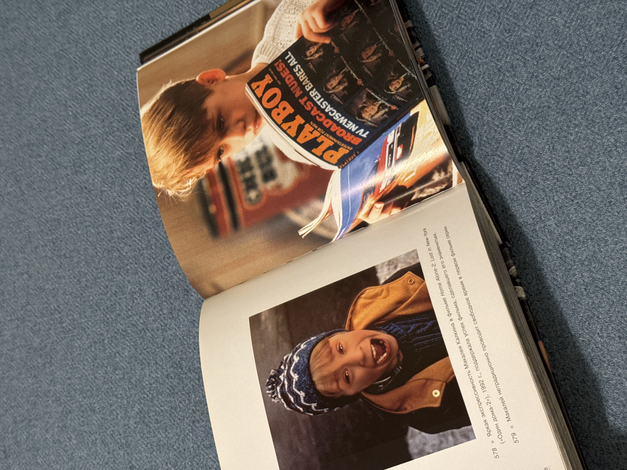 The image shows an open book resting on a blue denim-like surface. The left page features a picture of a young boy reading a Playboy magazine. The magazine cover has the headline "PLAYBOY" in large orange letters and the smaller text "BROADCAST NUDES! TV NEWSCASTER BARES ALL". The right page of the book shows a picture of Macaulay Culkin wearing a beanie and a jacket, screaming. Text in Russian is printed below the picture on the right page. The text and the photographs on the pages suggest the book is related to films or famous actors.