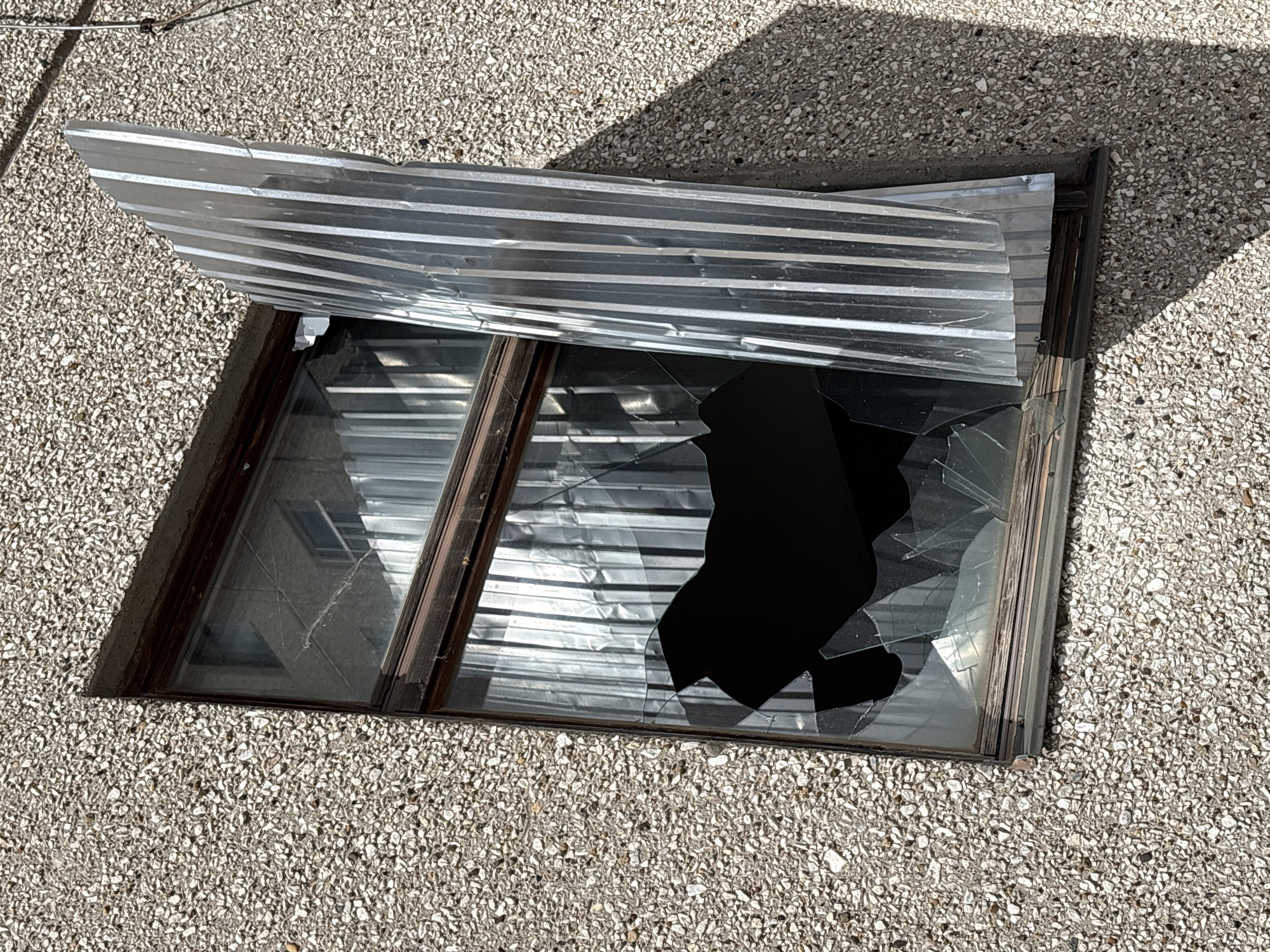 A basement window, partially covered by corrugated metal, is broken. The window is set into a light-colored, pebbled surface. Some glass shards are present, while a large portion of the glass is missing, revealing a dark interior. No people are visible. The location is not possible to determine from the given image.