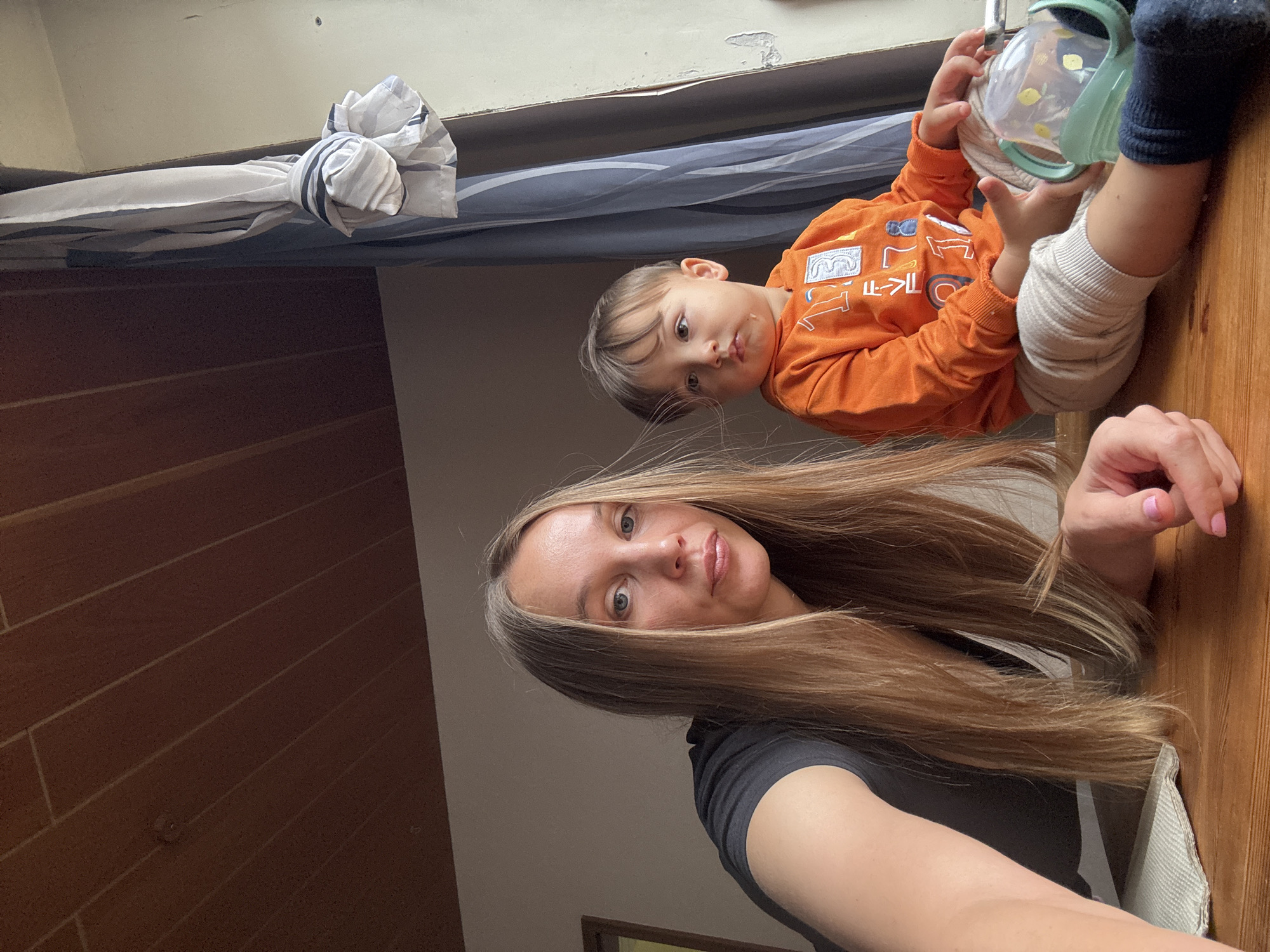 A woman and a young child are pictured indoors, likely in a residential setting. The woman, with long blonde hair, is wearing a black t-shirt and is positioned on the left side of the frame, looking towards the camera. Her right arm is extended, and her hand is resting on a wooden surface. The child, on the right, is wearing an orange long-sleeved shirt with graphics and light-colored pants with dark socks. The child is looking off to the side with a neutral expression and is holding a sippy cup with green handles. The background features a wooden-paneled ceiling, a curtain, and a wall. The scene appears to be lit by natural light coming from a window. There are no clear indicators of the specific city, Überherrn, Germany, within the image itself.