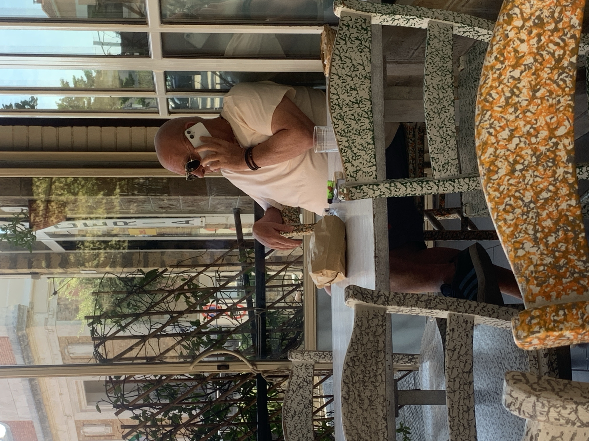 An outdoor seating area of a cafe or restaurant in San Quirico d'Orcia, Italy, is depicted. A bald man with sunglasses is seated at a table, holding a white smartphone to his ear and speaking on it. He is wearing a light-colored t-shirt and dark shorts. On the table are a brown paper-wrapped object, a glass of water, and cigarette packs. Several decorative chairs with white and patterned upholstery surround the table. The background shows a building with brickwork and large windows, reflecting the sky and surrounding greenery. A trellis with climbing plants is visible through a window.