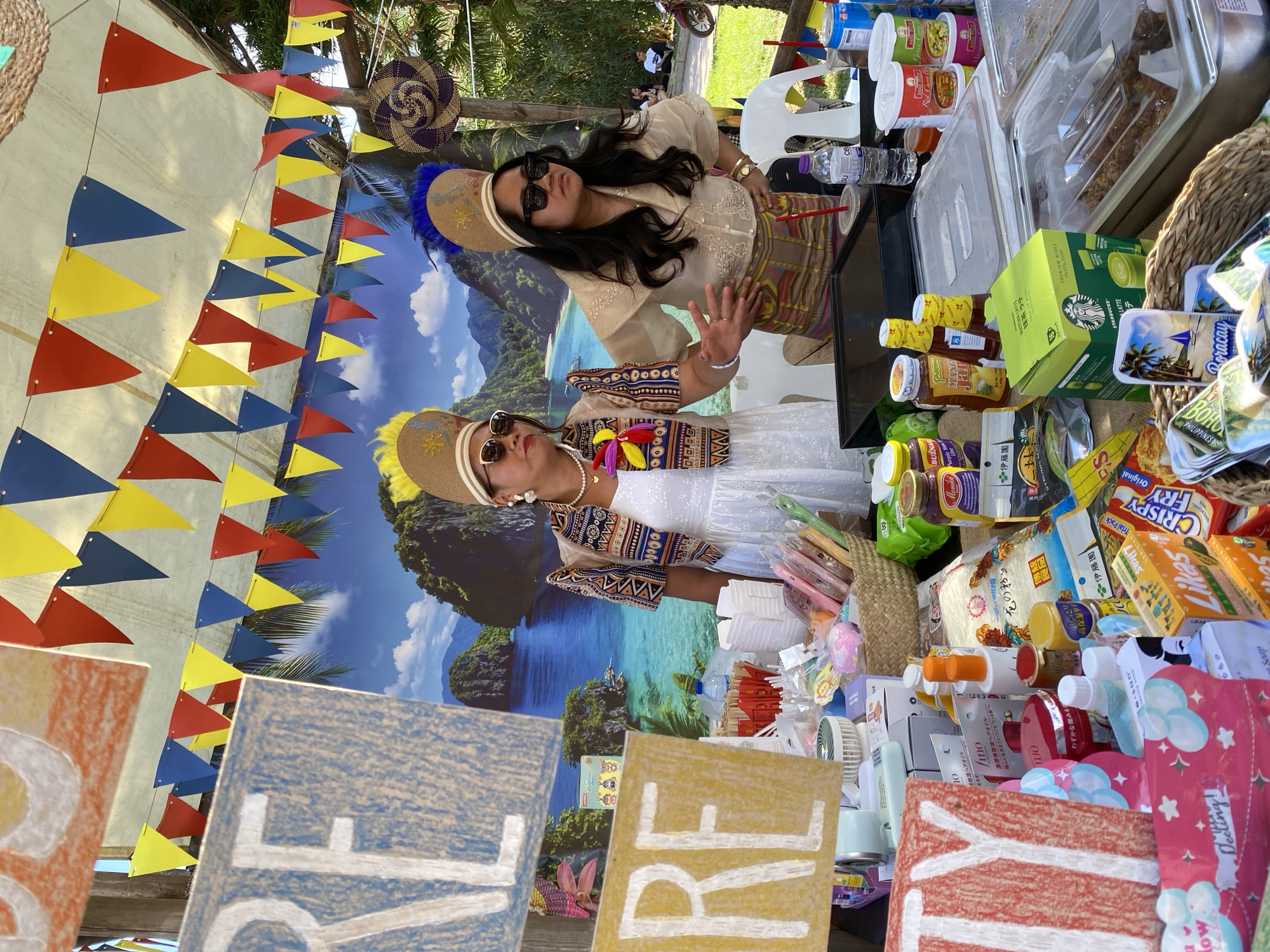 The image shows two women at an outdoor vendor stall with a backdrop depicting a tropical island scene, presumably Boracay, Philippines. Both women are wearing traditional Filipino attire and sunglasses. One woman is in a beige blouse and patterned skirt, waving her hand, while the other is in a white dress with a patterned jacket. The stall is laden with various items, including food containers, drinks, snacks, personal care products, and souvenirs. Signs with painted letters reading "RE" and "LOVE" are visible in the foreground. Strings of red, blue, and yellow triangular flags are strung above the scene. A woven circular object hangs on the side.
The location in the city of Alanya, Turkey cannot be determined from the image.