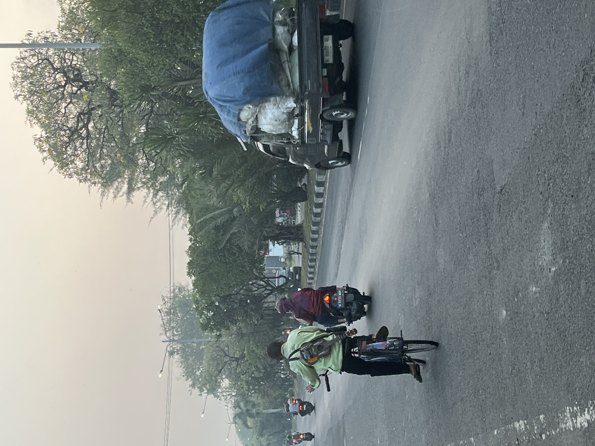 In Ngaban, Indonesia, a cyclist wearing a light green jacket and a backpack with a guitar attached rides on the right side of a paved road. Behind him, a person on a motorbike wearing a maroon hijab and jacket is also traveling in the same direction. A small truck, covered with a blue tarp, is driving ahead on the right lane. The scene is set against a hazy sky, with trees lining the road on the right side. Multiple other motorbikes are visible in the distance on the left side of the road.