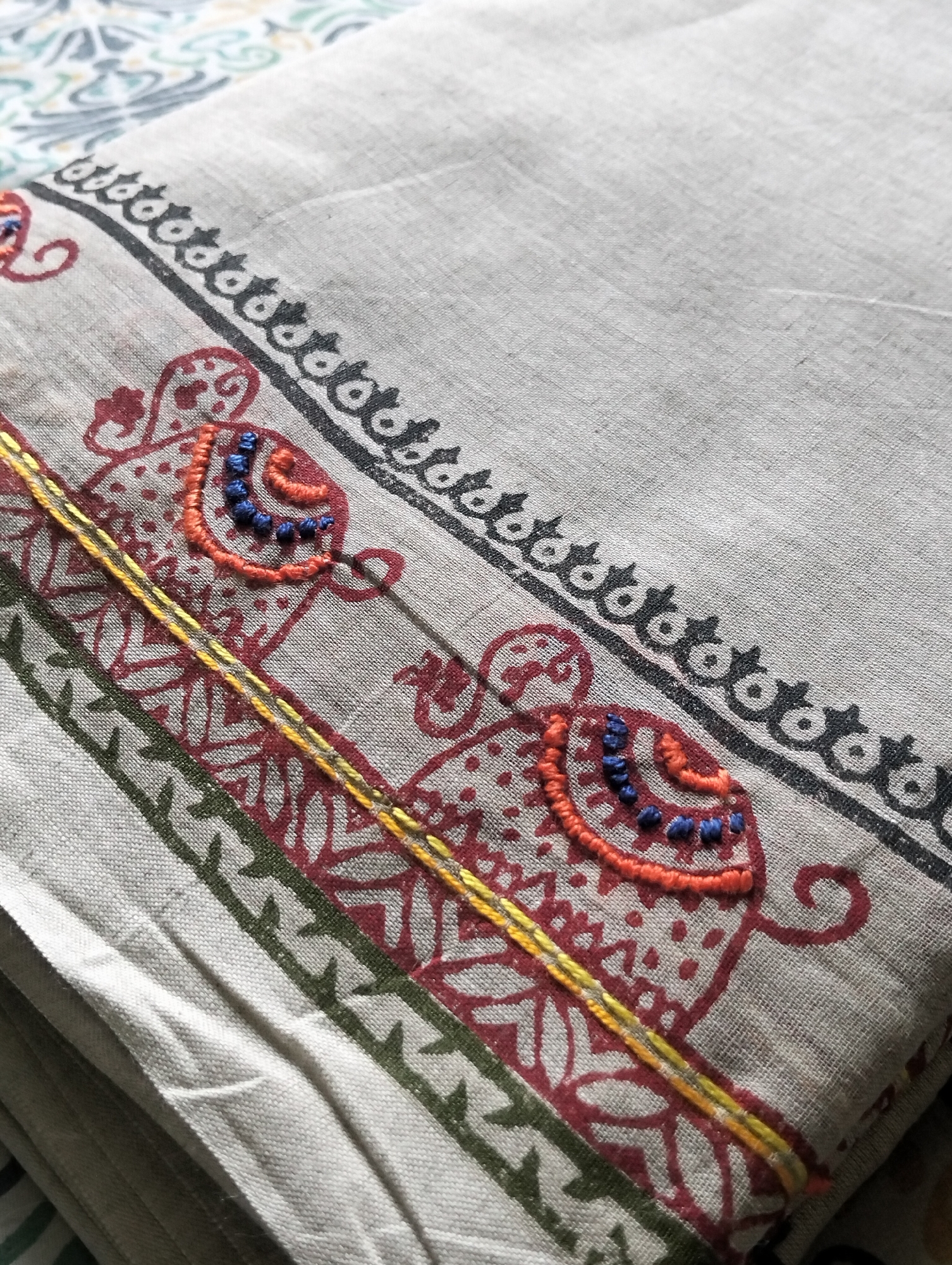 A close-up shot shows a folded piece of fabric with printed and embroidered designs. The fabric is a light gray or beige color with a visible woven texture. Along the edge, there's a repeating black pattern that looks like a series of curved shapes. Below that, there are red (or possibly maroon) floral or paisley-like designs with orange and blue embroidery on top. A yellow embroidered line runs parallel to the red pattern. Further down, there's a band of green with a simple repeating pattern.
Without additional context, it is impossible to determine the exact location within the city of Dhaka, Bangladesh, where this image was taken. There are no people visible in the image.