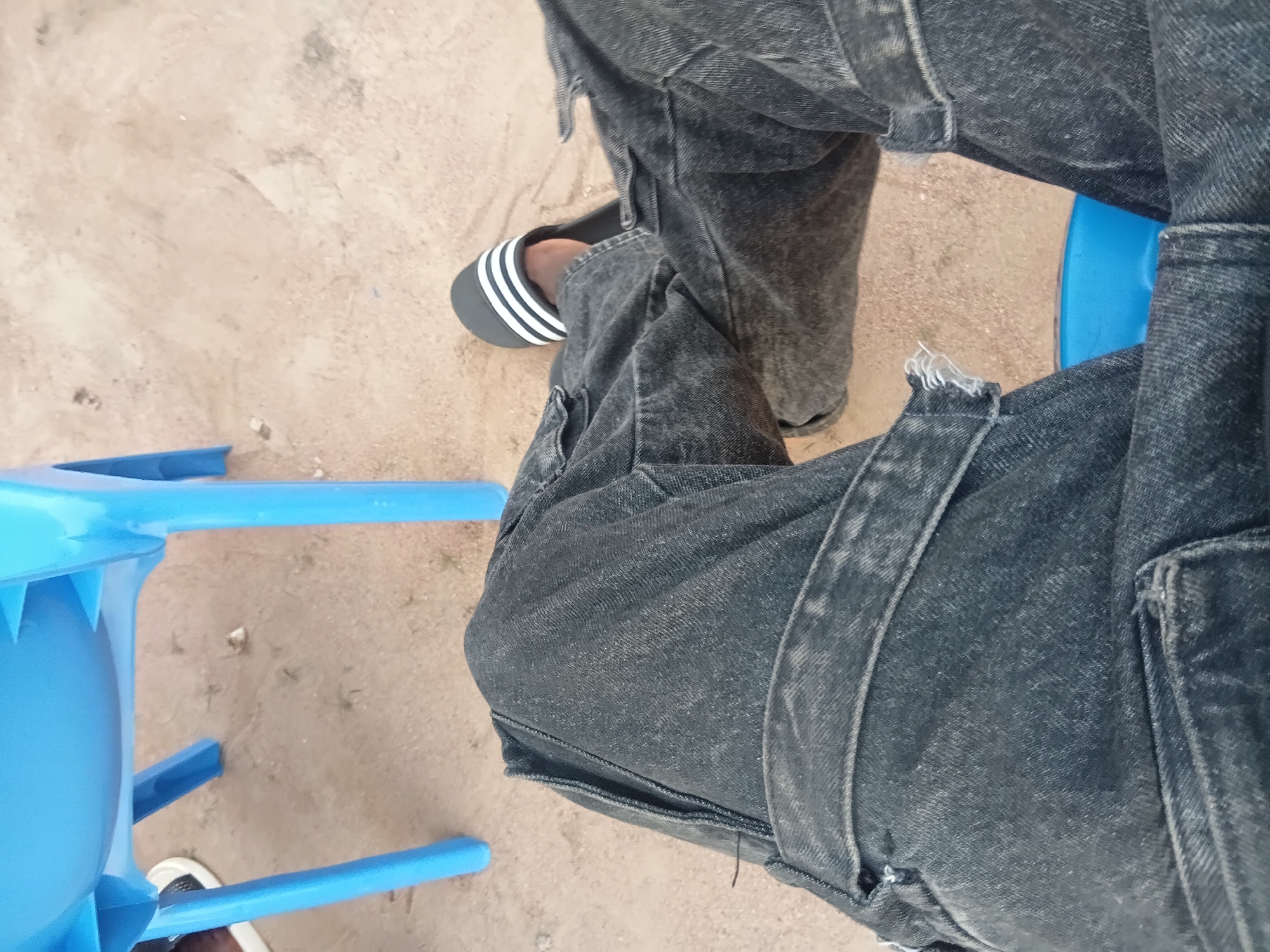 The image shows a person's legs from the knee down, wearing dark denim cargo pants and a black Adidas slide sandal with white stripes. The person is seated on an unseen surface, with their legs positioned over a bright blue plastic object, possibly a bucket or stool. The background consists of light brown, sandy ground with scattered debris. The overall scene appears to be outdoors, in a casual setting. There is no identifiable information within the image that would specify a location in Jalingo, Nigeria.