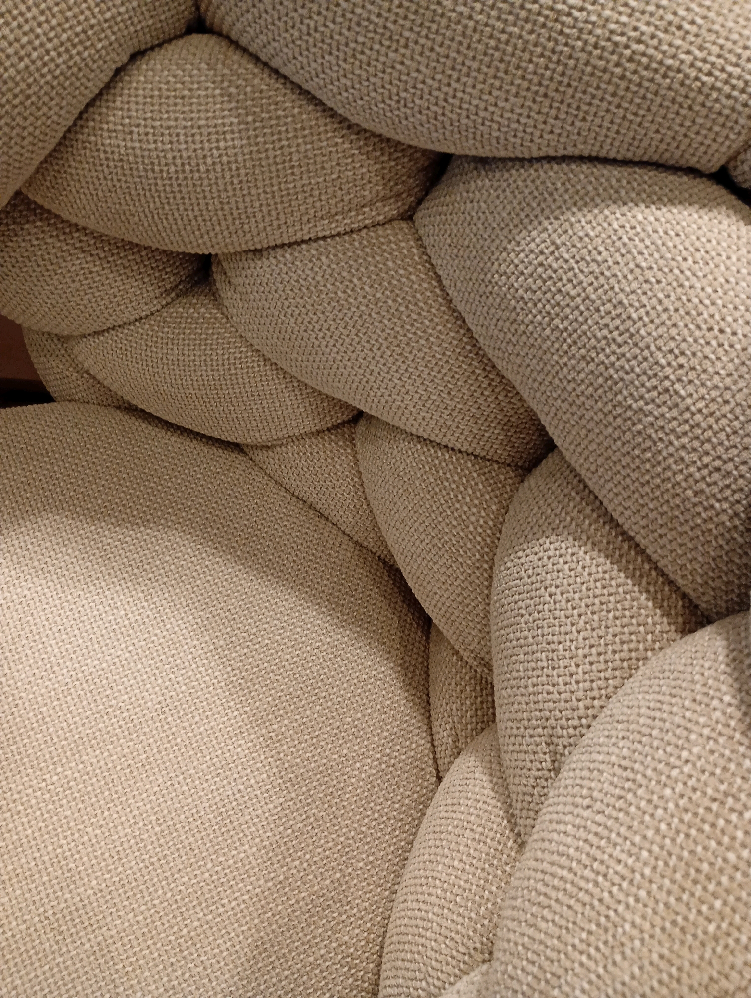 The image shows a close-up, overhead view of a braided, beige-colored fabric, likely upholstery on a piece of furniture. The texture of the fabric is visible, with a woven pattern. The lighting creates soft shadows, emphasizing the rounded, tubular forms of the braid. There are no people, actions, or environmental details that suggest a specific city or location like Bad Sachsa, Germany. The image is a still life focused solely on the texture and form of the braided fabric.