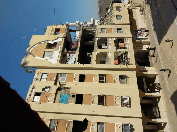 The image depicts a multi-story residential building in Khan Yunis, Palestinian Territory, exhibiting significant structural damage under a clear blue sky. The upper floors, particularly the top-right section, are extensively damaged, with large portions of the facade and roof missing, exposing interior rooms. Windows are broken or absent, and balconies show wear and tear, with some displaying hanging laundry. The building's exterior is light brown with decorative arched window surrounds and vertical column accents.
At street level, below the building's overhang, a silver sedan is parked. A group of approximately five individuals, including adults and children, are gathered in the shaded area beneath the building. On the adjacent asphalt street, two adults, one dressed in dark attire, and two children are observed walking. To the right of the main structure, an adjacent building section is reduced to a pile of rubble. A mosque minaret is partially visible in the background, further right. A mural is present on a wall next to the rubble.
