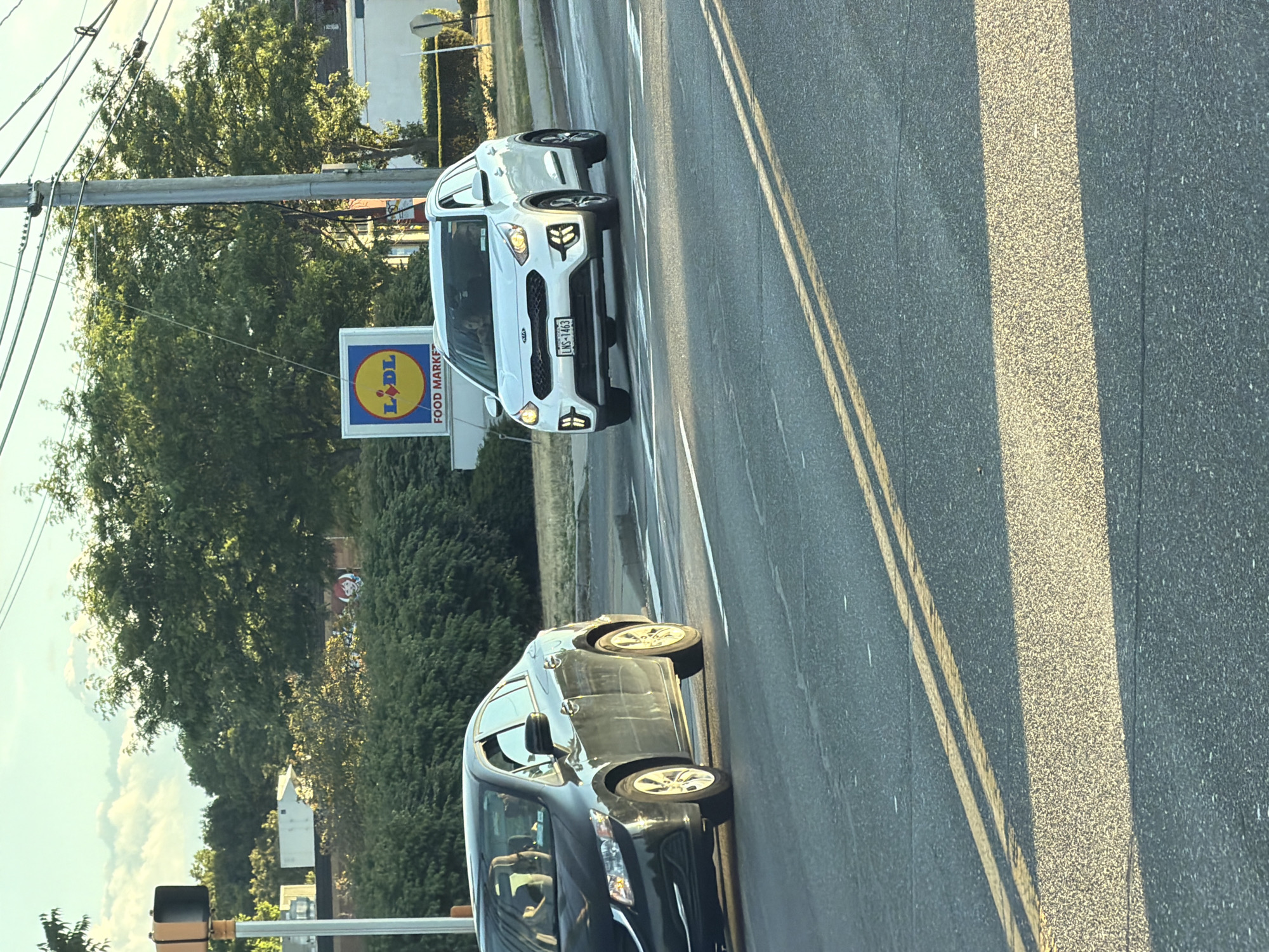 In Riverhead, New York, on an overcast day with dappled sunlight, two vehicles are visible on a road. In the foreground, a dark-colored sedan, possibly a Honda Civic, is in the left lane, its headlights illuminated. In the right lane, a white Kia Niro SUV is traveling forward, its headlights also on. The Kia's license plate reads "LNS-1463." Behind the vehicles, a sign for "Lidl Food Market" is prominent, featuring the distinctive yellow and blue logo. Trees and bushes line the background, with power lines and poles overhead. The road has double yellow lines marking the lanes.