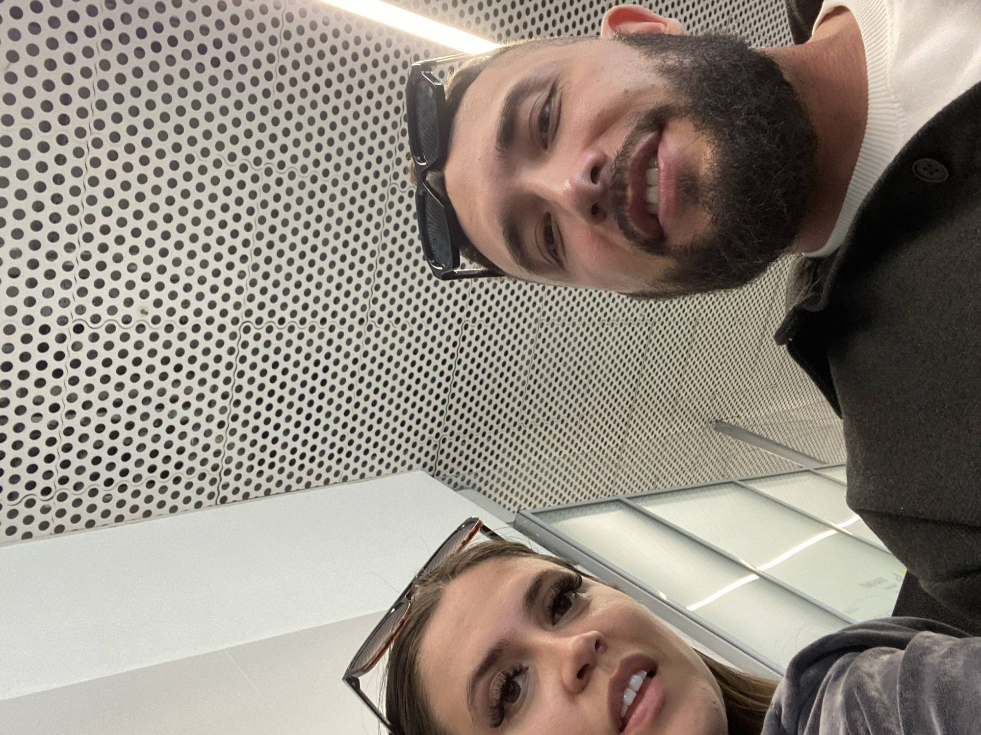 In this close-up selfie, a man and a woman are looking at the camera. The woman, on the left, has brown hair and is wearing a grey velour top. She has large false eyelashes and is wearing sunglasses on top of her head. The man, on the right, has a beard and is wearing a dark jacket over a white sweater. He is also wearing sunglasses on top of his head. They appear to be sitting next to each other, possibly in a public waiting area. The background consists of a textured ceiling with a pattern of small holes, and some white panels with lights are visible to the left. The scene does not provide any specific visual cues to indicate the location in the city of Новое Мартьяново, Russia.