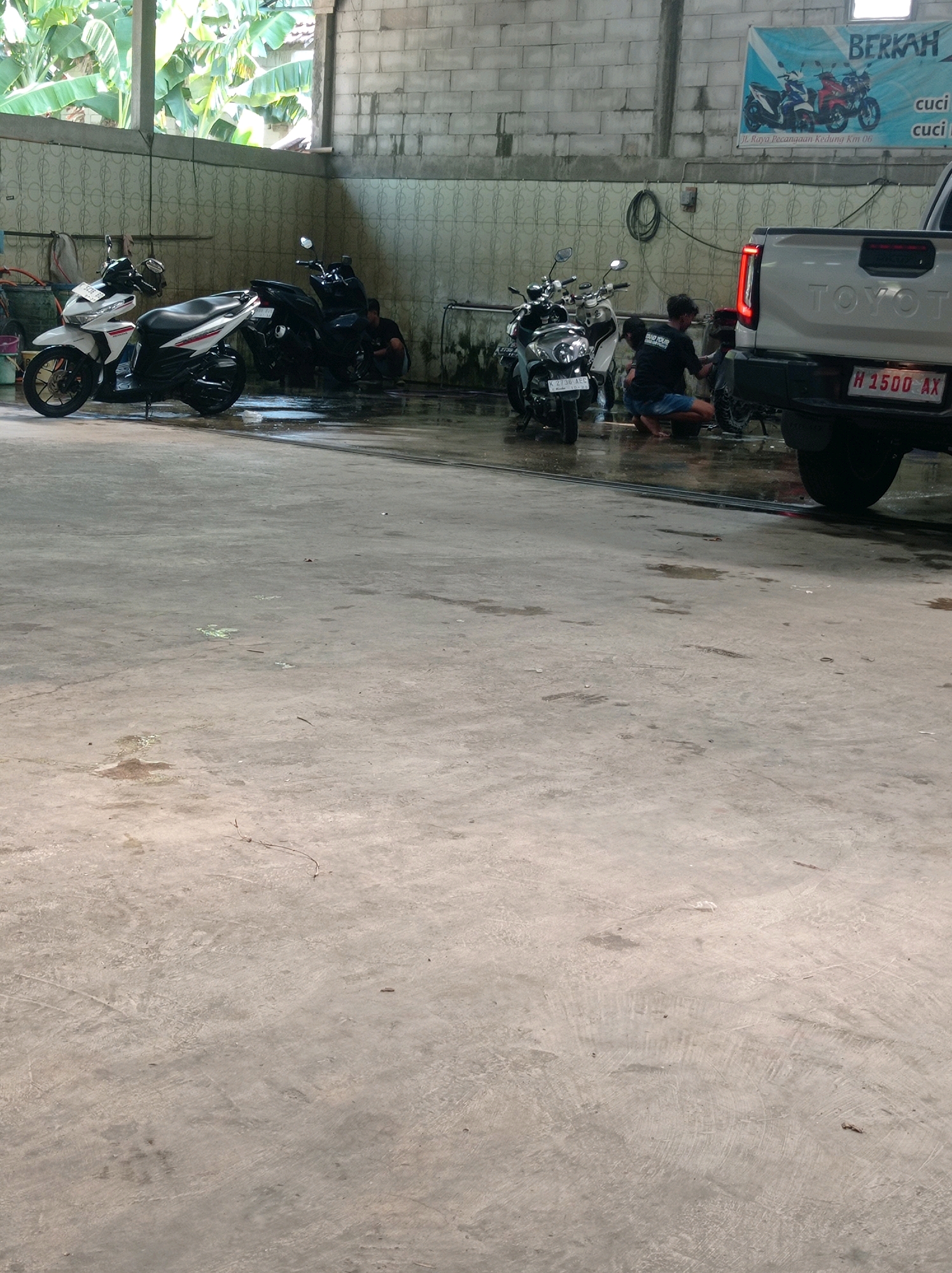 This image depicts a car and motorcycle wash facility located in Kedungmalang, Indonesia. The scene is set inside a covered structure with a concrete floor that appears wet from washing activities. To the left, two motorcycles are parked, one white and one black. In the background, a white Toyota pickup truck is visible, with its rear end facing the camera. Two individuals are actively engaged in washing a motorcycle near the truck. One person is crouched down, appearing to be cleaning the rear wheel, while the other stands nearby, holding what might be a cleaning tool. The walls of the structure are made of cinder blocks and white tiles, and a banner advertising "BERKAH CUCI" (blessed wash) with images of motorcycles is displayed on the wall. Lush green foliage is visible through an open section of the structure, suggesting an outdoor or semi-outdoor setting. The overall impression is a busy, functional car wash operation.