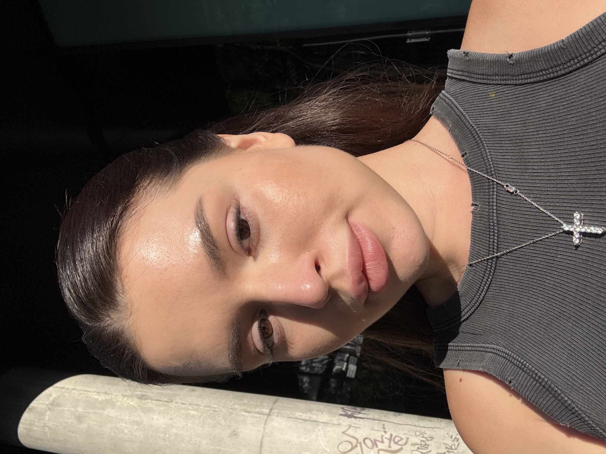A close-up, waist-up, eye-level shot features a young woman with dark hair and brown eyes facing forward, her head tilted slightly to her right. She is wearing a dark grey ribbed tank top with distressed shoulder straps and a silver necklace with a jeweled cross pendant. The woman's skin appears dewy and reflects the sunlight.
The background is divided into distinct elements. To the left, a large, light-colored concrete pillar with graffiti is visible. To the right, a dark, out-of-focus background suggests an urban environment, possibly a building facade or foliage. The lighting indicates it is daytime, with strong sunlight casting shadows and highlighting the woman's features. The image does not provide specific indicators of its location within Prague, Czechia.