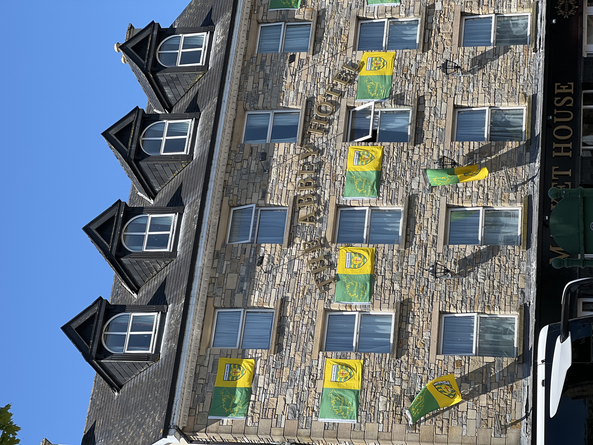The image shows a stone-clad building facade featuring multiple stories with several windows and dormer windows. The building is adorned with numerous green and yellow flags, some displaying the crest of Donegal and text that reads "Dún Na Ngall" and "Donegal." The building's signage prominently reads "THE ABBEY HOTEL." Below the hotel, a sign for "MARKET HOUSE" is visible. A black and white vehicle, possibly a bus or van, is partially visible in the foreground at the bottom of the image. The sky is clear and blue. The scene is likely set in the city of Donegal, Ireland, given the prominent display of Donegal-themed flags and the building's name. There are no people or animals visible in the image. The overall impression is one of a building decorated for a local event or celebration, possibly related to sports given the county colors.