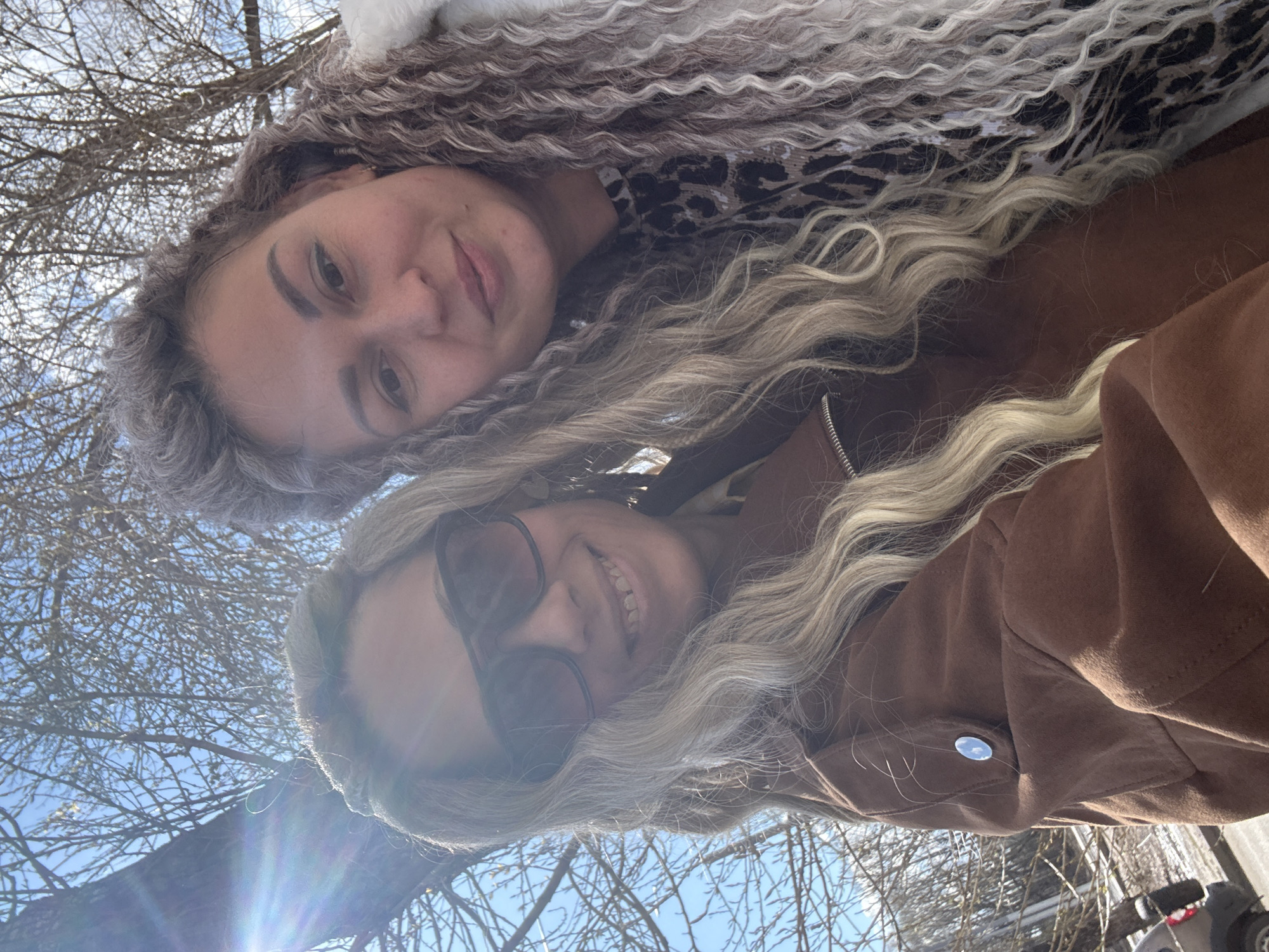 Two women are posing for a selfie. The woman at the top has curly grey and brown hair and is wearing a patterned top. The woman at the bottom has blonde hair and is wearing sunglasses and a brown jacket. Behind them is a clear blue sky and bare tree branches. A building and vehicle are visible in the lower right corner. The location may be Шебунино, Russia, based on a search term provided.