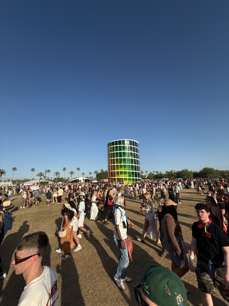 A wide-angle shot captures a daytime scene at what appears to be a large outdoor event in Indio, California. A dense crowd of people occupies a grassy field. The architecture of the "Spectra" art installation stands as a central point in the background. Palm trees line the horizon. The sky is cloudless and blue. Individuals are diverse in appearance and clothing style, suggesting a festival or gathering. Shadows of people cast on the grass are elongated, indicating a relatively low sun angle.