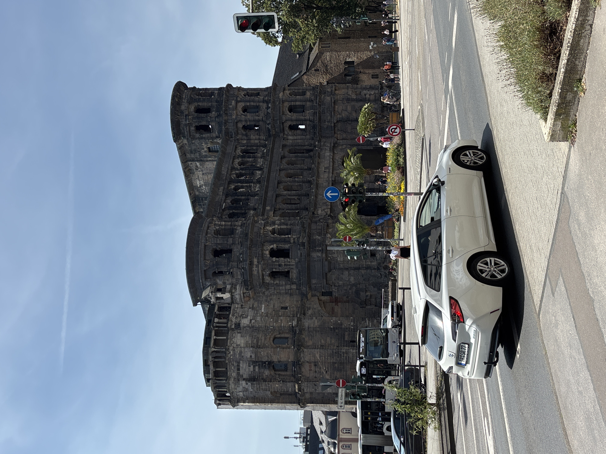 The image shows the Porta Nigra, a Roman city gate in Trier, Germany, on a sunny day. A white station wagon is driving on the street in the foreground, with its rear license plate visible, reading "TR SR 77". In the background, several people are walking near the historic structure and a bus is stopped at an intersection. Traffic lights and street signs are also visible. The sky is clear blue with a faint contrail.