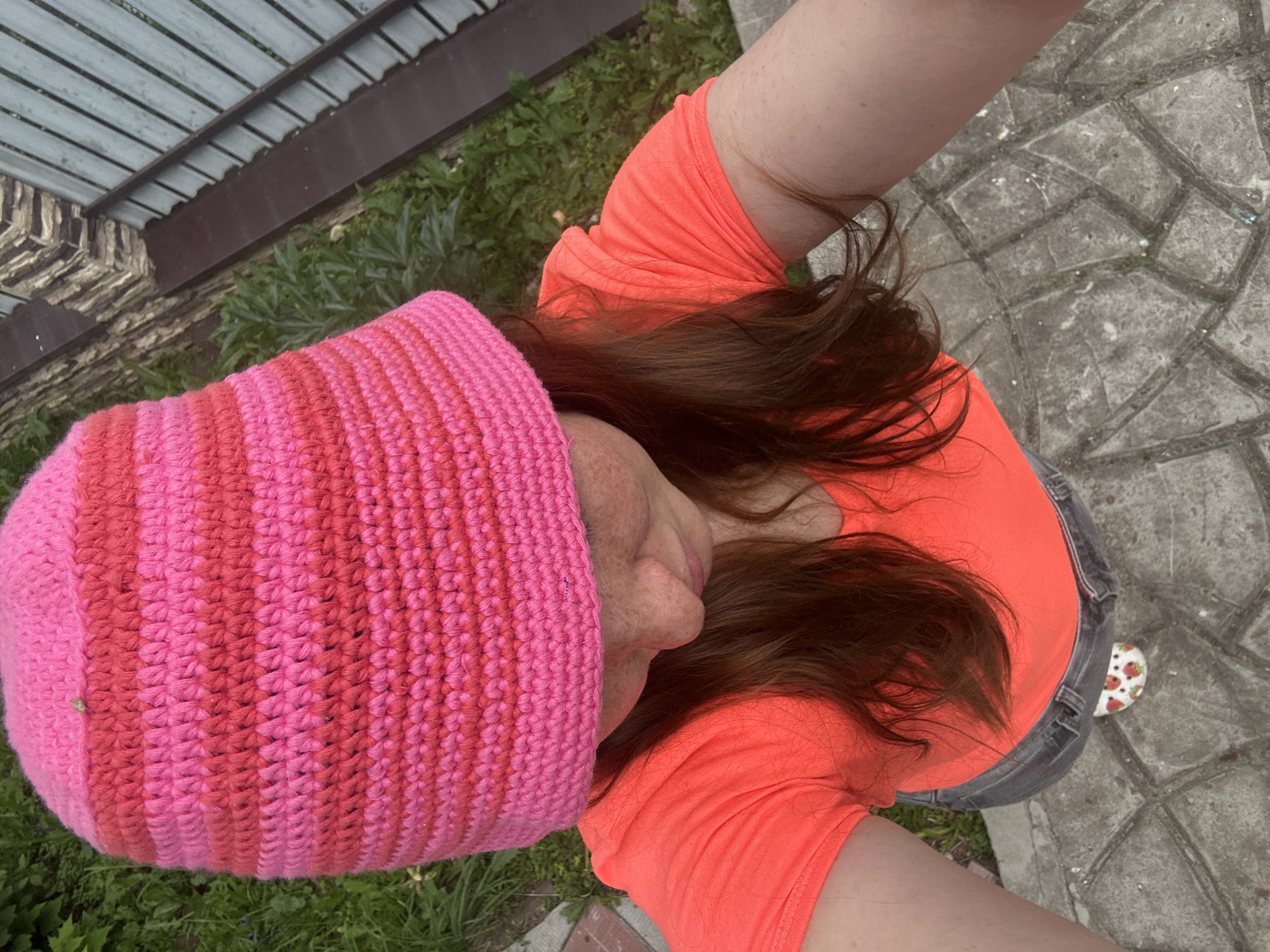 The image shows a first-person perspective of a person wearing a bright pink and coral crocheted beanie and an orange t-shirt. Their long, brown hair is visible, and they have freckles on their face. They are wearing grey denim jeans. The person is standing on a paved walkway made of interlocking grey stones. To the left of the walkway, there is a patch of green grass and a fence. The fence appears to be made of wooden slats on top of a metal structure. The overall scene suggests an outdoor setting, possibly a yard or a public area. The lighting is natural, indicating it is daytime, and there are no strong shadows, suggesting it might be an overcast day or the sun is high in the sky. The image appears to be a selfie taken from a high angle, looking down at the person and their surroundings. The focus is on the person and their attire, with the background providing context. The person's head is tilted, and their face is partially visible, suggesting a casual pose. A small object with a strawberry pattern is visible near the person's foot on the paved ground. No visible text is present in the image.