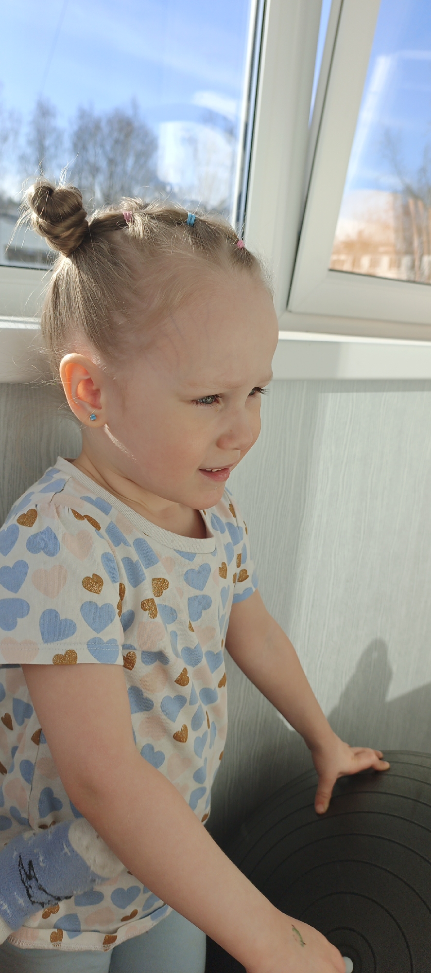 A young, light-haired child, likely a girl, is pictured indoors next to a window, looking down and to her right with a gentle smile. Her blonde hair is styled in a top bun, secured with small colorful hair ties (pink and blue). She wears a small blue stud earring in her left ear. Her light-colored t-shirt is patterned with blue, light pink, and golden glitter hearts. Light blue pants are visible below her shirt, and a blue sock with a white design (possibly a donkey character) is visible on her left arm. A small green temporary tattoo is on her right wrist.
The child's right hand rests on a large, dark grey or black exercise ball. Behind her is a textured, light-colored wall. A large window fills the upper background, revealing a clear blue sky and bare trees in the distance, indicating a bright daytime setting, possibly early spring or late autumn in Chaikovsky, Russia. Strong sunlight streams through the window, illuminating the child's face and hair. The overall mood is one of contentment and quiet engagement. No visible text is present in the image.