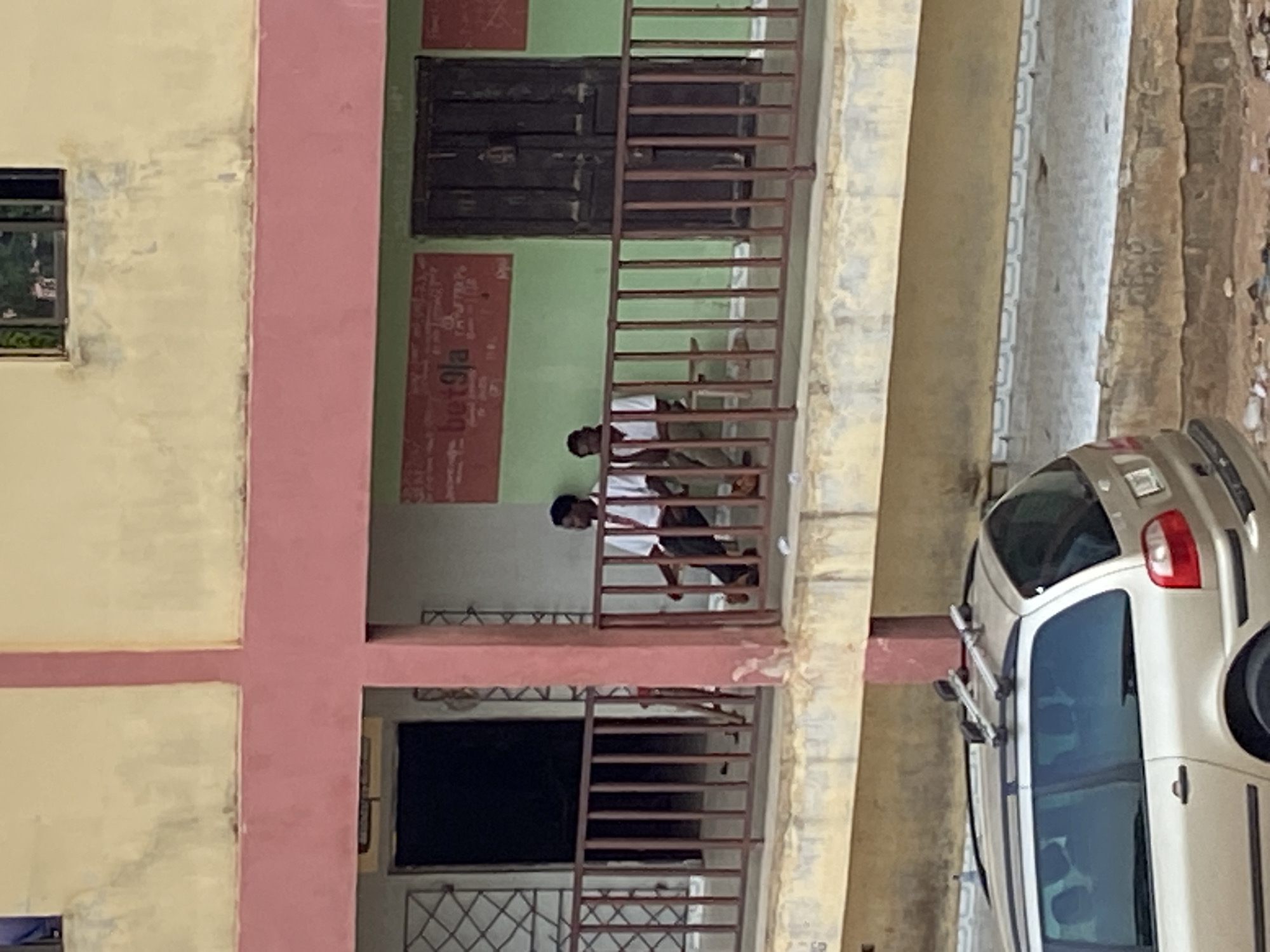 This image depicts an exterior view of a multi-story building in Mararaba, Nigeria, captured during daylight hours. The building features cream-colored walls contrasted with distinct pink vertical and horizontal architectural elements. An upper floor includes sections painted green, behind which dark wooden doors or shutters are visible.
On a balcony or open walkway on this upper floor, two young males are present. They are dressed in light-colored shirts and dark trousers, standing near a brown metal railing that extends horizontally across the frame. Their activity appears to be simply standing or observing their surroundings.
To the right of the building, on what seems to be a street or alley, a light-colored (possibly silver or beige) car, resembling a hatchback or small SUV, is visible. The car is equipped with a roof rack and is parked or moving alongside the building. The ground surrounding the car shows signs of wear and minor debris.
A visible detail on the green wall behind the two individuals is the word "bet9ja" printed in white, vertically oriented text. The scene conveys a glimpse into an everyday urban or semi-urban environment.
