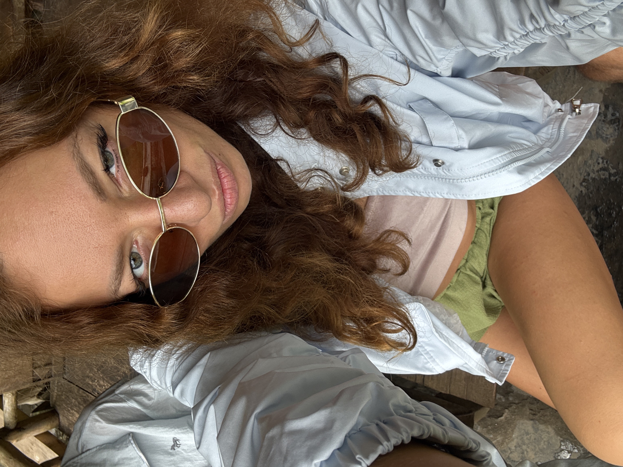A woman with curly brown hair wearing sunglasses is posing for a picture. She is wearing a light blue, button-down shirt, a light pink top, and green shorts. The woman has fair skin and green eyes. The background consists of a weathered wooden surface and a textured, grey stone-like surface. The image is angled. The picture may have been taken in Yên Minh Commune, Vietnam.