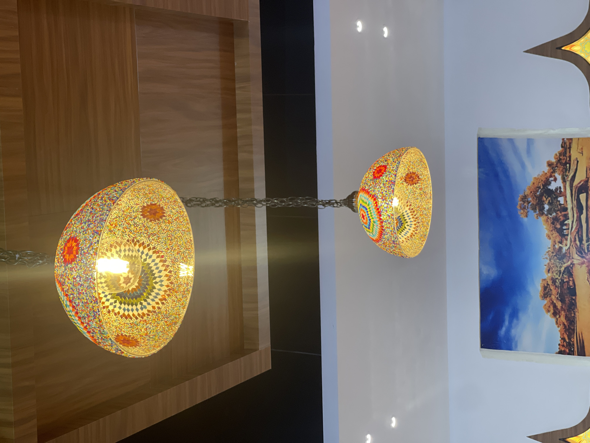 A photograph taken indoors shows two hanging mosaic-style lamps with chains suspended from the ceiling. One lamp hangs in front of a wooden panel wall, while the other is positioned in front of a white wall adjacent to a framed picture depicting a sky and land scene with trees. The photo was taken in Chongqing High-tech Zone, China.